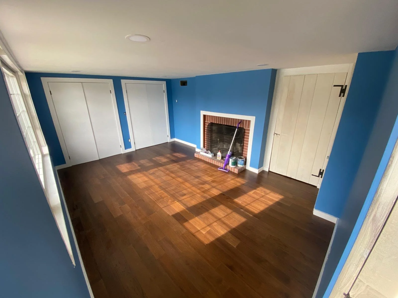 Empty living room with blue walls, a fireplace, and hardwood floors, featuring three closed white doors, a mop and cleaning supplies by the fireplace, and a window on the left.
