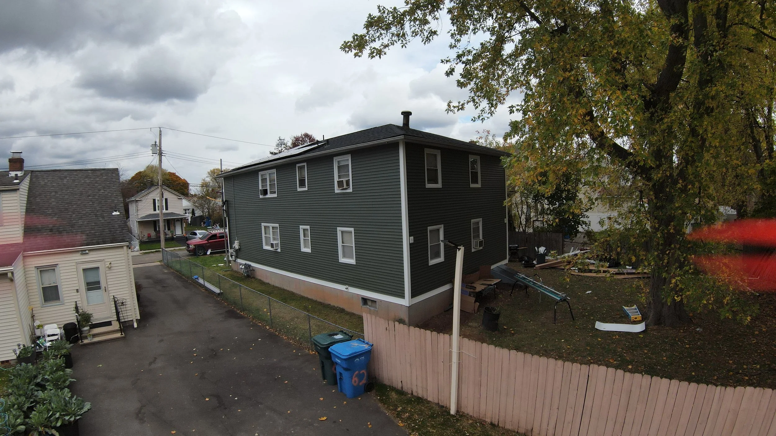 A residential backyard with a large tree, a grey two-story house, a fenced yard, and two trash cans on the driveway. Overcast sky with gray clouds.