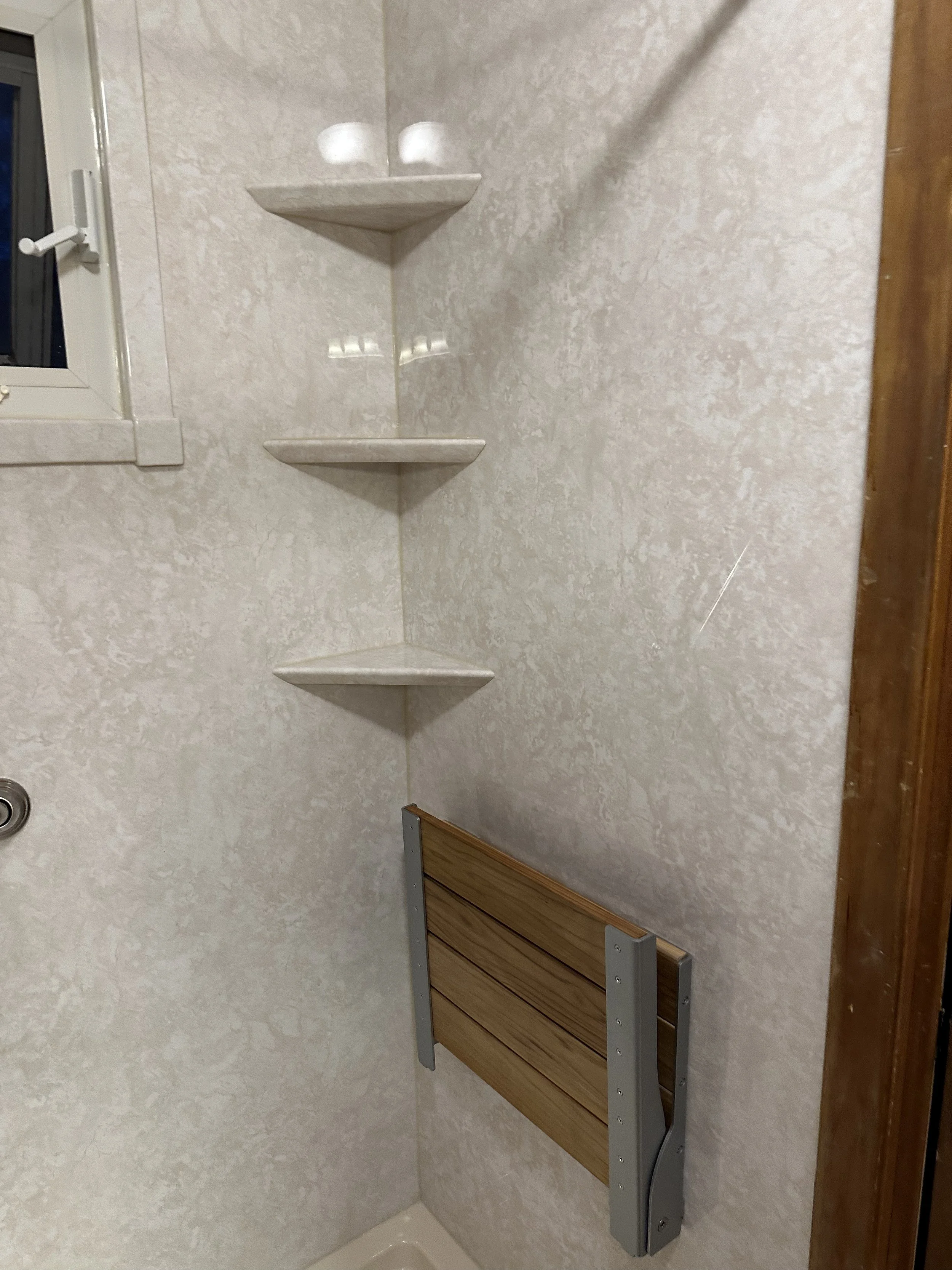 Corner of a shower with three floating corner shelves, a small window with a latch, and a wooden fold-down seat.