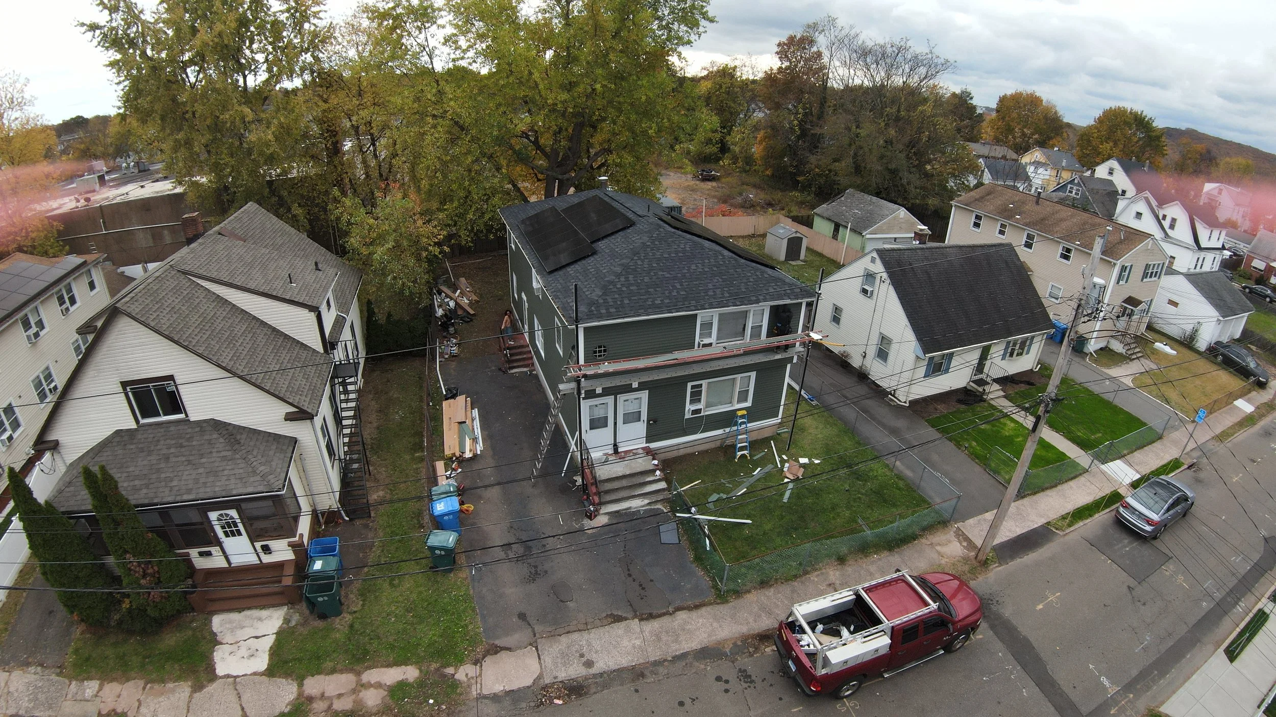 An aerial view of a residential neighborhood with a house under renovation, scaffolding around it, and solar panels on the roof. Surrounding houses are visible, some with solar panels, lawns, and cars parked on the street.