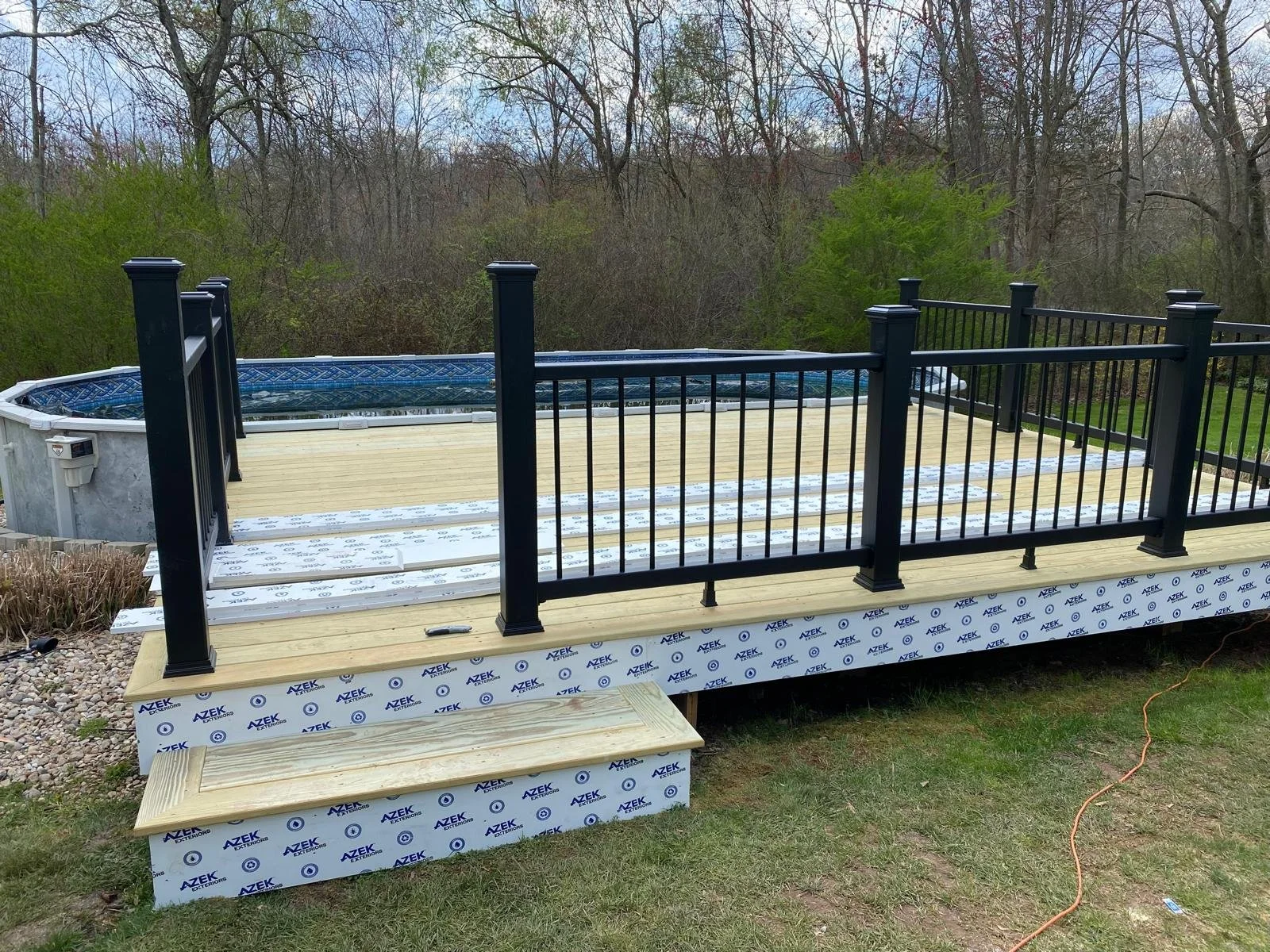 Partially constructed wooden deck with black metal railing and an above-ground pool in a backyard, with trees in the background.