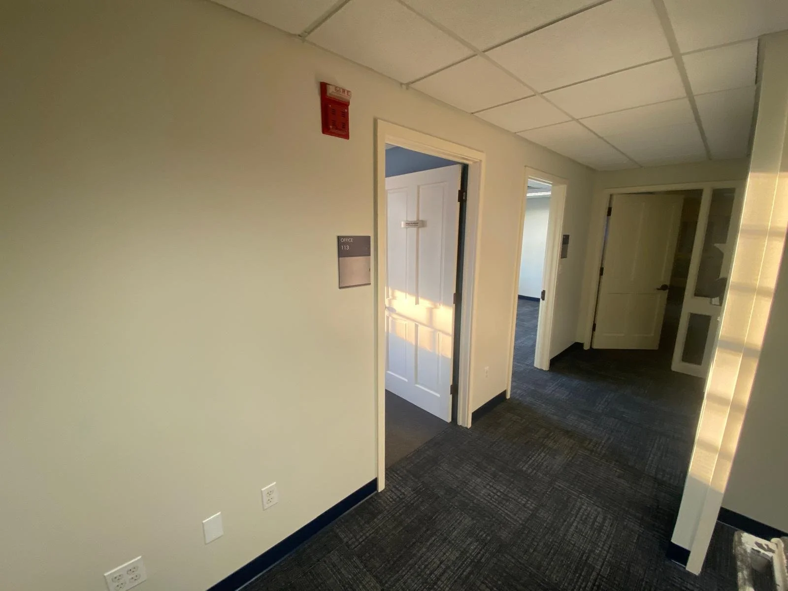 Interior hallway of an office building with closed doors, a fire alarm on the wall, and office signs.