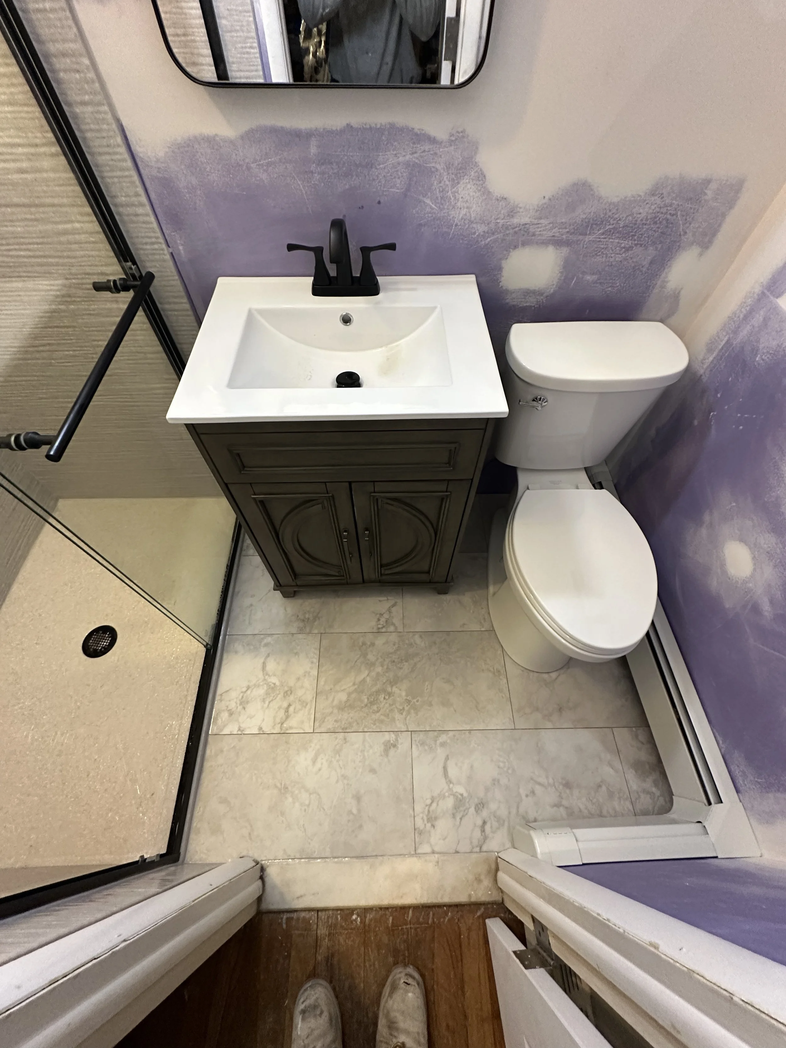 Small bathroom with a white sink and a black faucet, a mirror above the sink, a toilet, a purple and white textured wall, and part of a shower with a glass door.