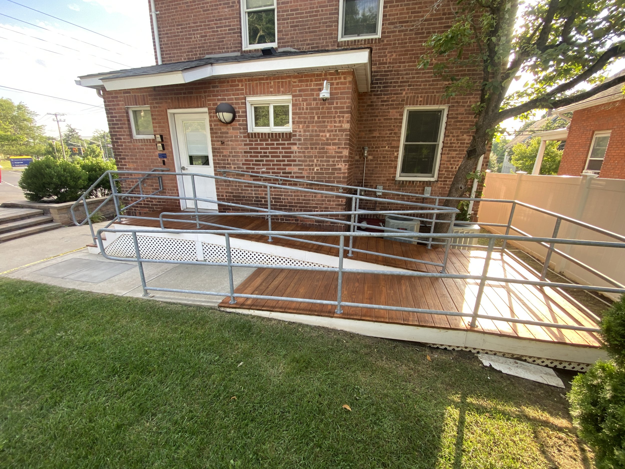 A house with a brick exterior featuring a small wooden porch with a ramp and metal railings, with steps to the side, surrounded by greenery and a fence.