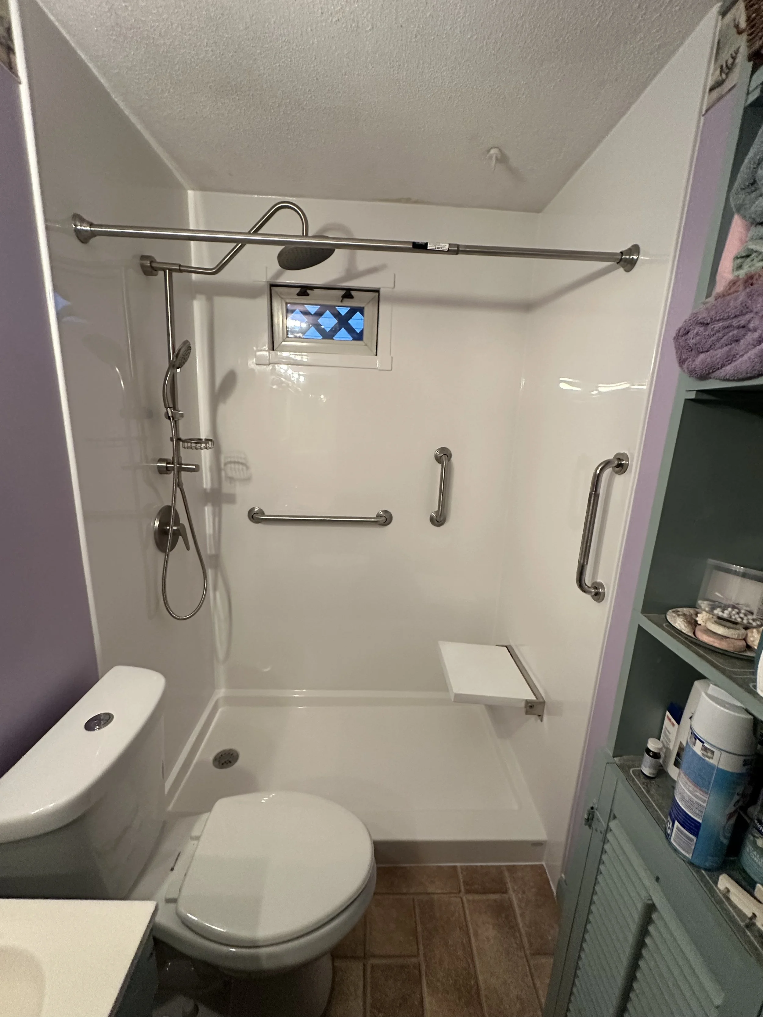 Bathroom with a walk-in shower featuring grab bars, a small fold-down seat, a handheld showerhead, and a showerhead mounted on the ceiling. There is a small window above the shower, a toilet, and shelves with toiletries on the right side.