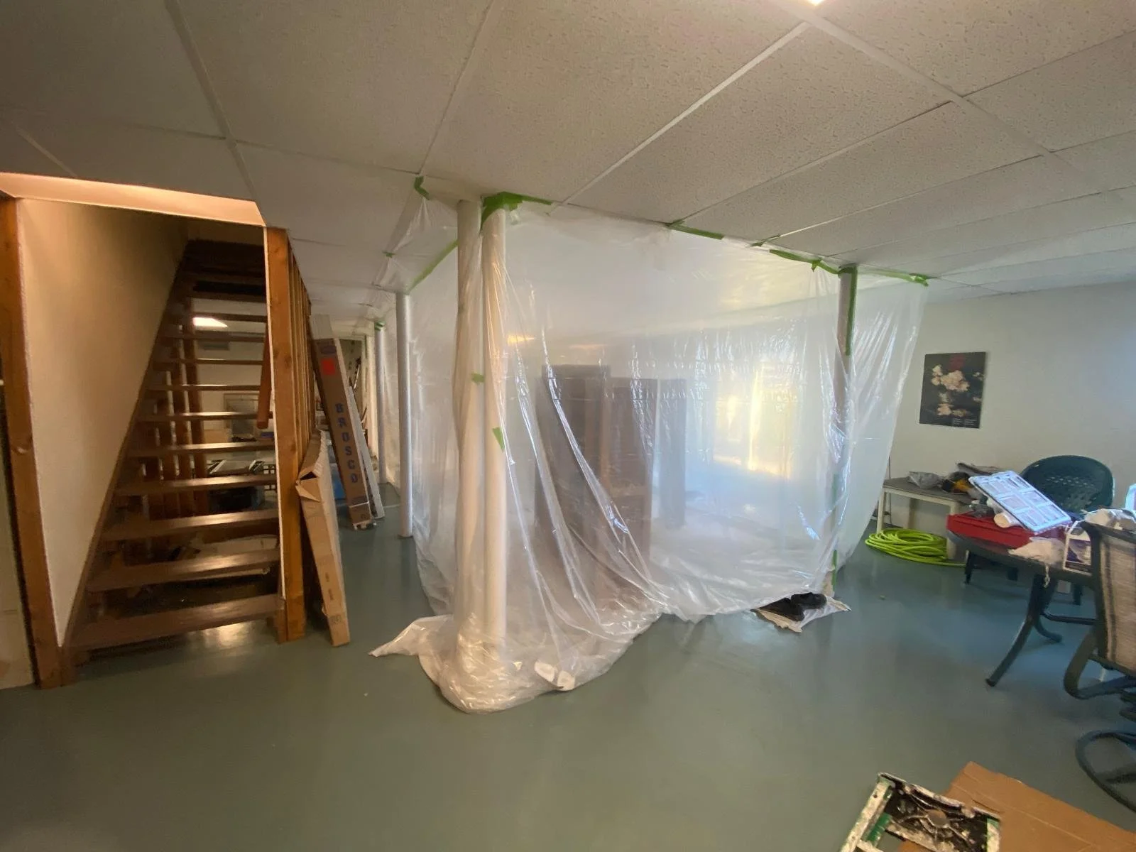 A basement under renovation with plastic sheeting hanging from the ceiling, a wooden staircase on the left, and various tools and furniture scattered around.