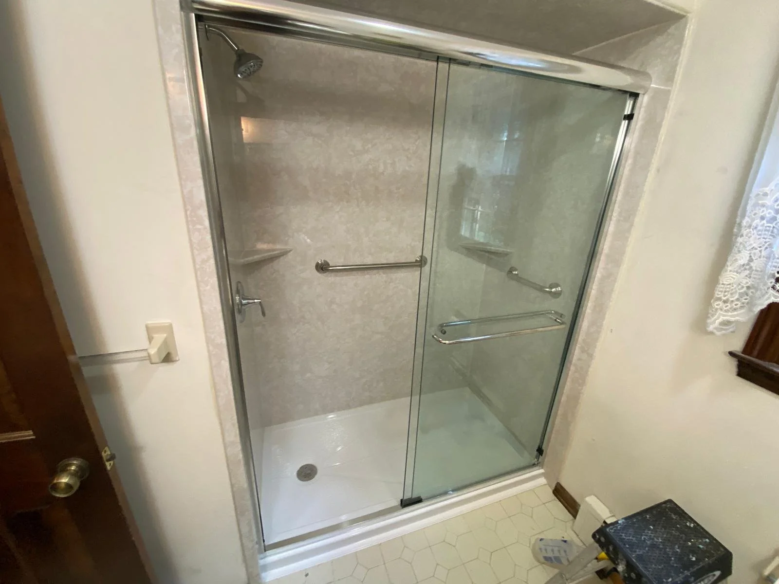A glass shower enclosure with silver frames and two sliding doors, silver grab bars, and a showerhead, in a bathroom with beige walls and hexagon-shaped white floor tiles.