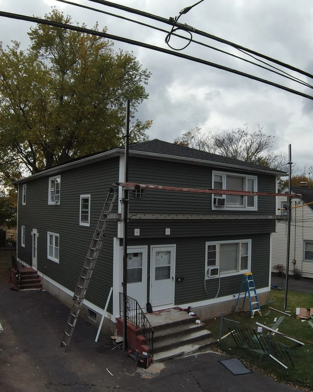 A two-story house under construction or maintenance with ladders and scaffolding, surrounded by tools and equipment, and an overcast sky.