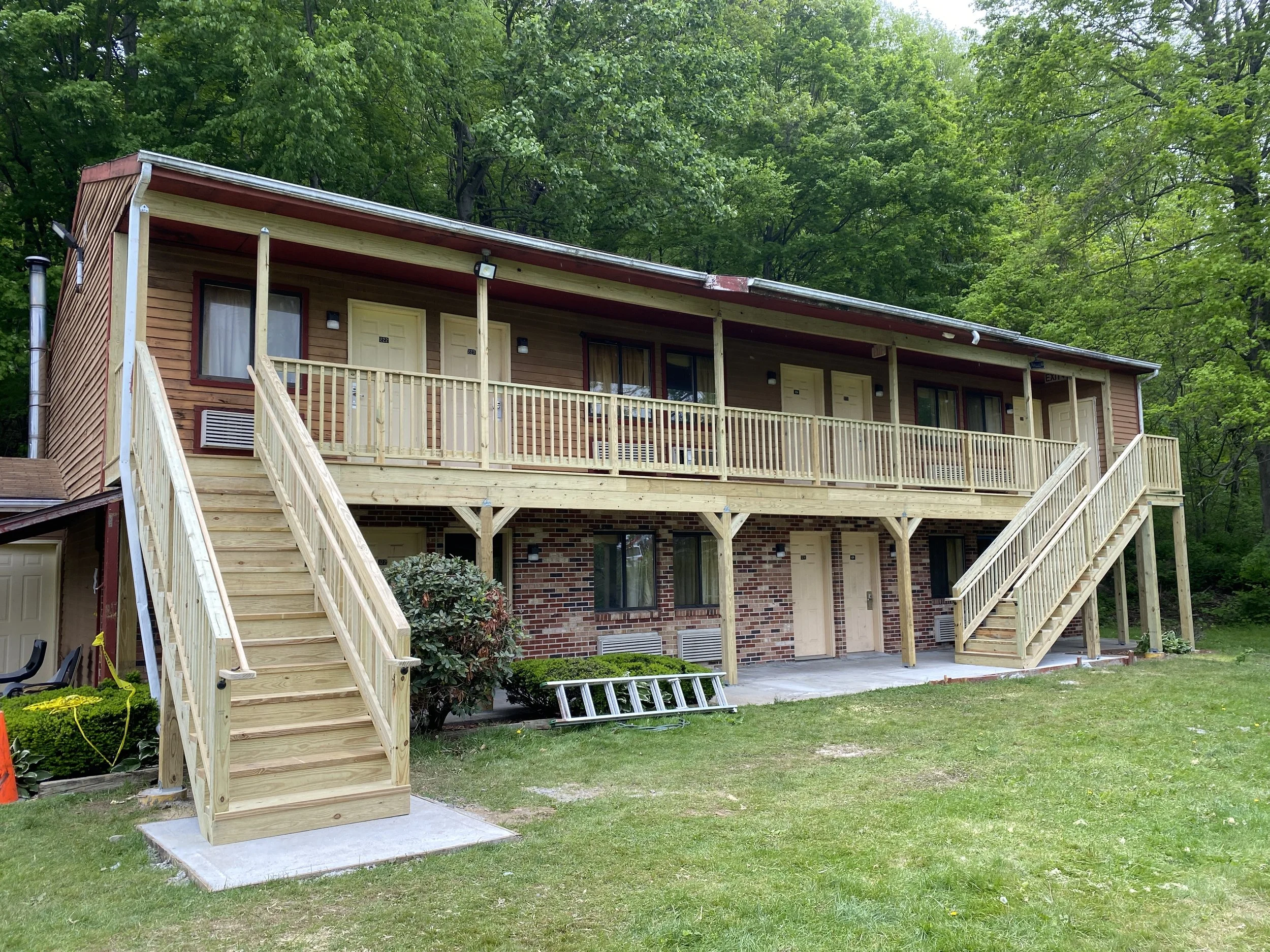 A two-story apartment building with balconies, stairs on both sides, brick and wood exterior, surrounded by green trees and grass.