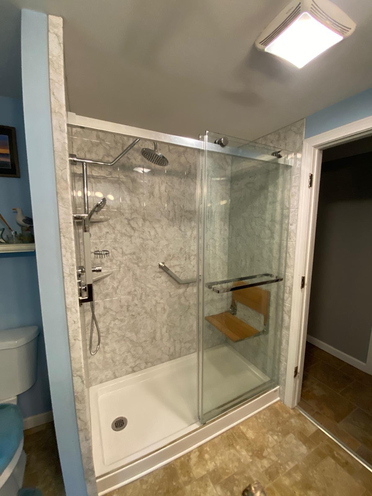 A bathroom shower with a glass door, marble walls, a showerhead, a handheld shower, and a wooden seat. The shower area has a white base and is next to a doorway.
