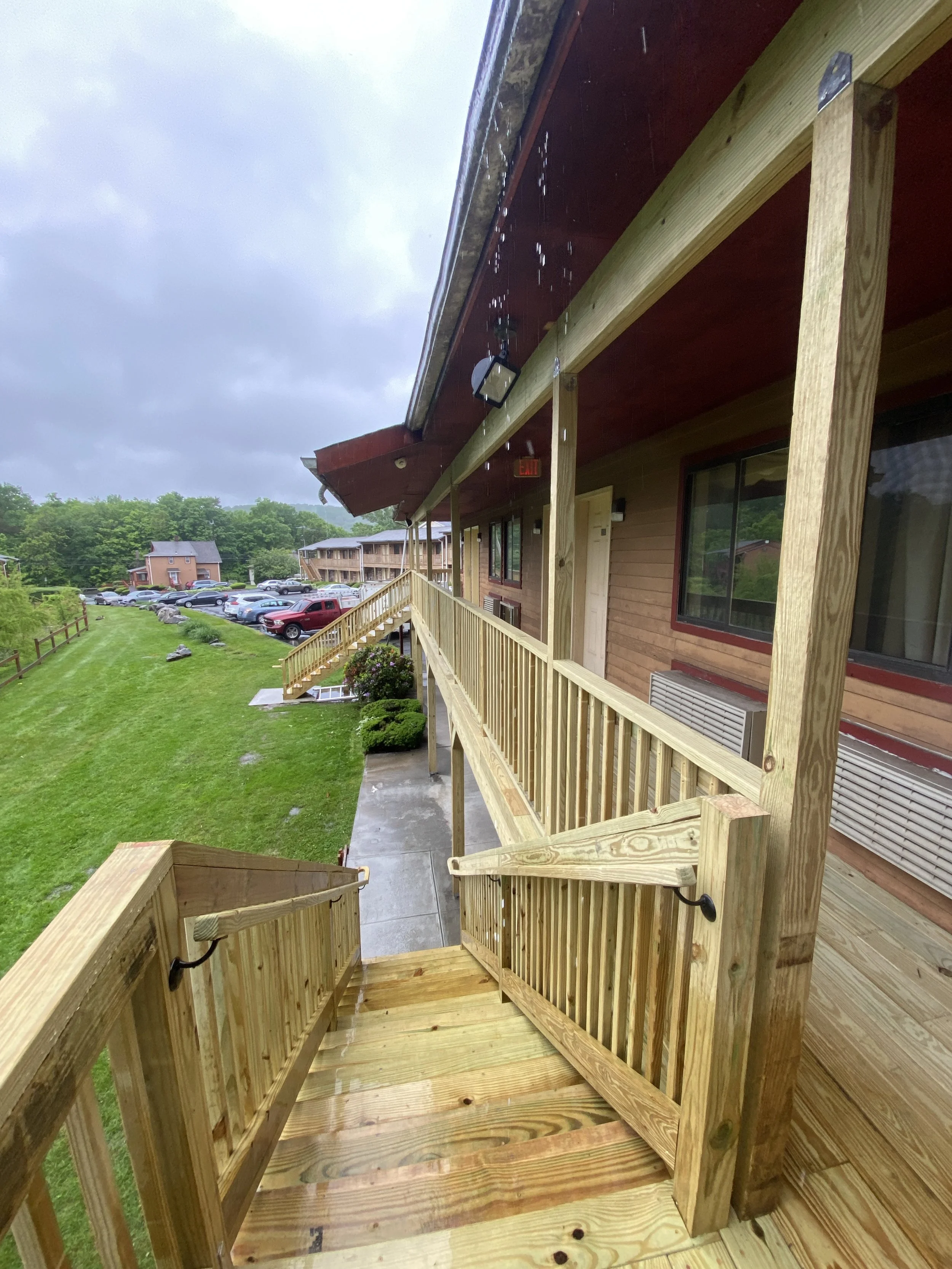 View of a wooden balcony with railing outside a building, overlooking a grassy area, parking lot with cars, and other buildings under cloudy sky.