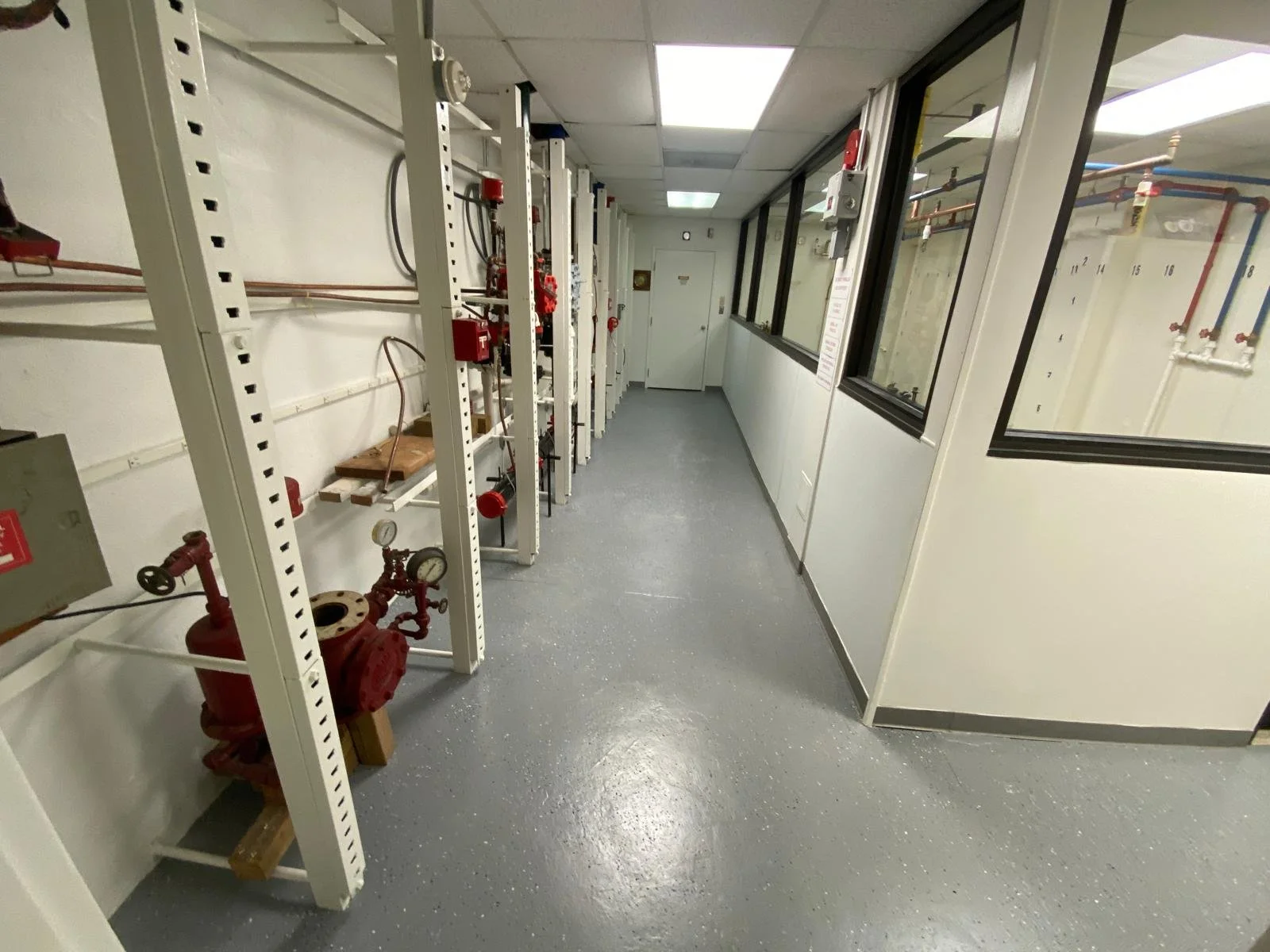 A hallway in a building with equipment and fire safety devices on the left wall and windows on the right wall, showing an industrial or maintenance area.