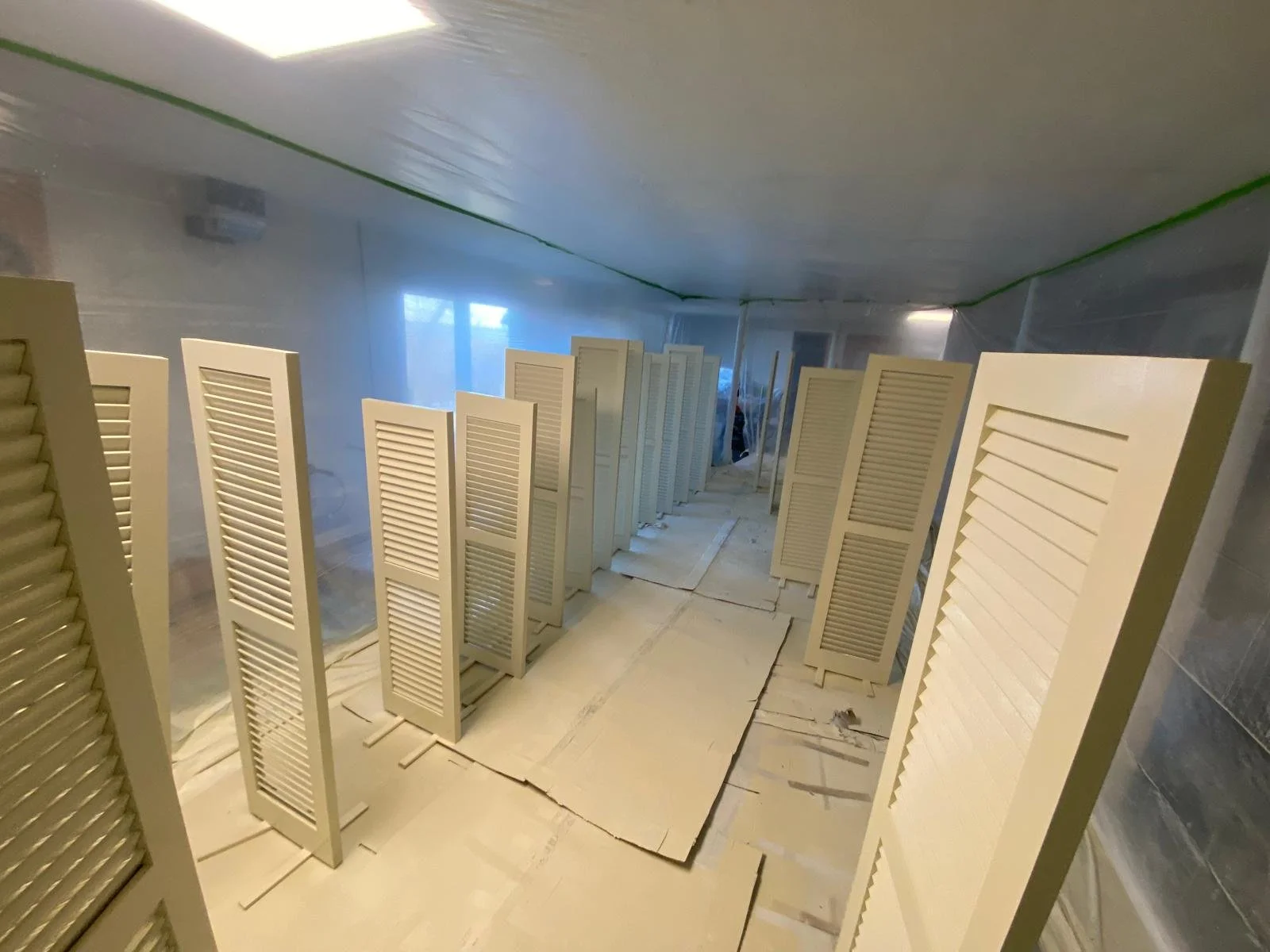 Rows of beige painted wooden louvered shutters in a room under renovation, with plastic sheeting on the walls and ceiling, and cardboard on the floor.