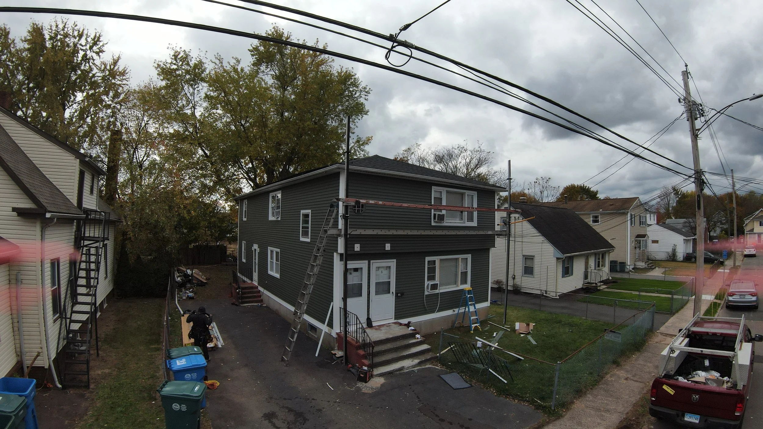 Construction workers repairing a house with scaffolding, ladders, and tools outside on a cloudy day in a neighborhood.