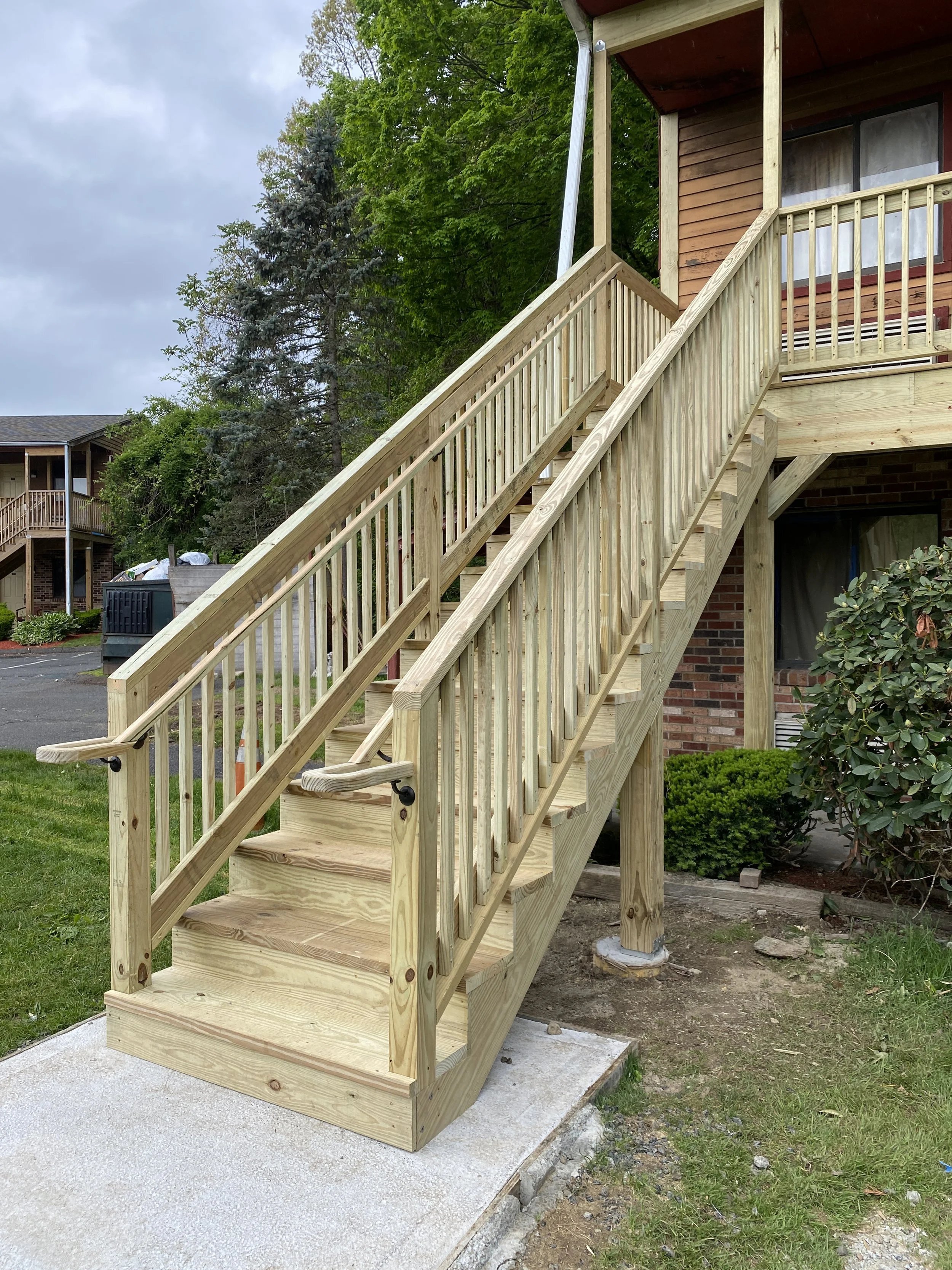 Newly built wooden staircase with handrails leading up to a balcony of a brick and wood house, with green trees and cloudy sky in the background.