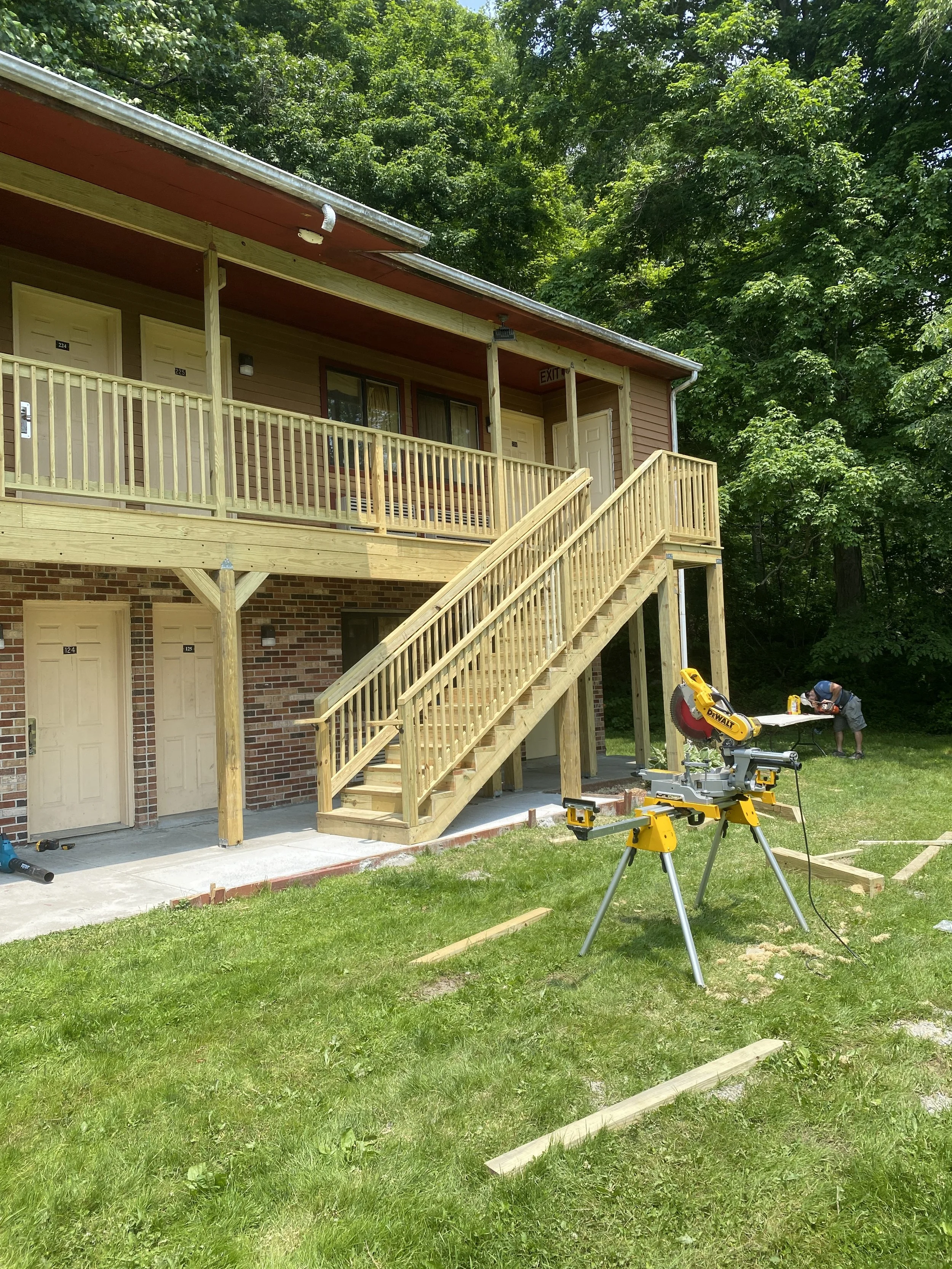 Construction scene of a new wooden staircase outside an apartment building, with a worker in the background. The building has a brick lower level and a wooden upper level with a balcony. Tools and materials are on the ground.