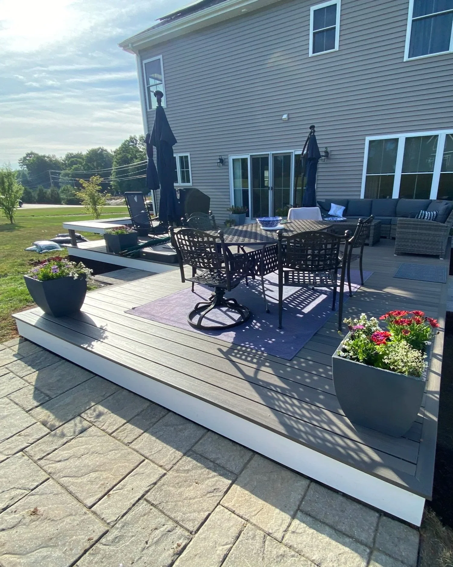 A backyard patio with outdoor furniture, including a round table, chairs, a sectional sofa, and umbrellas, surrounded by potted flowers in planters.