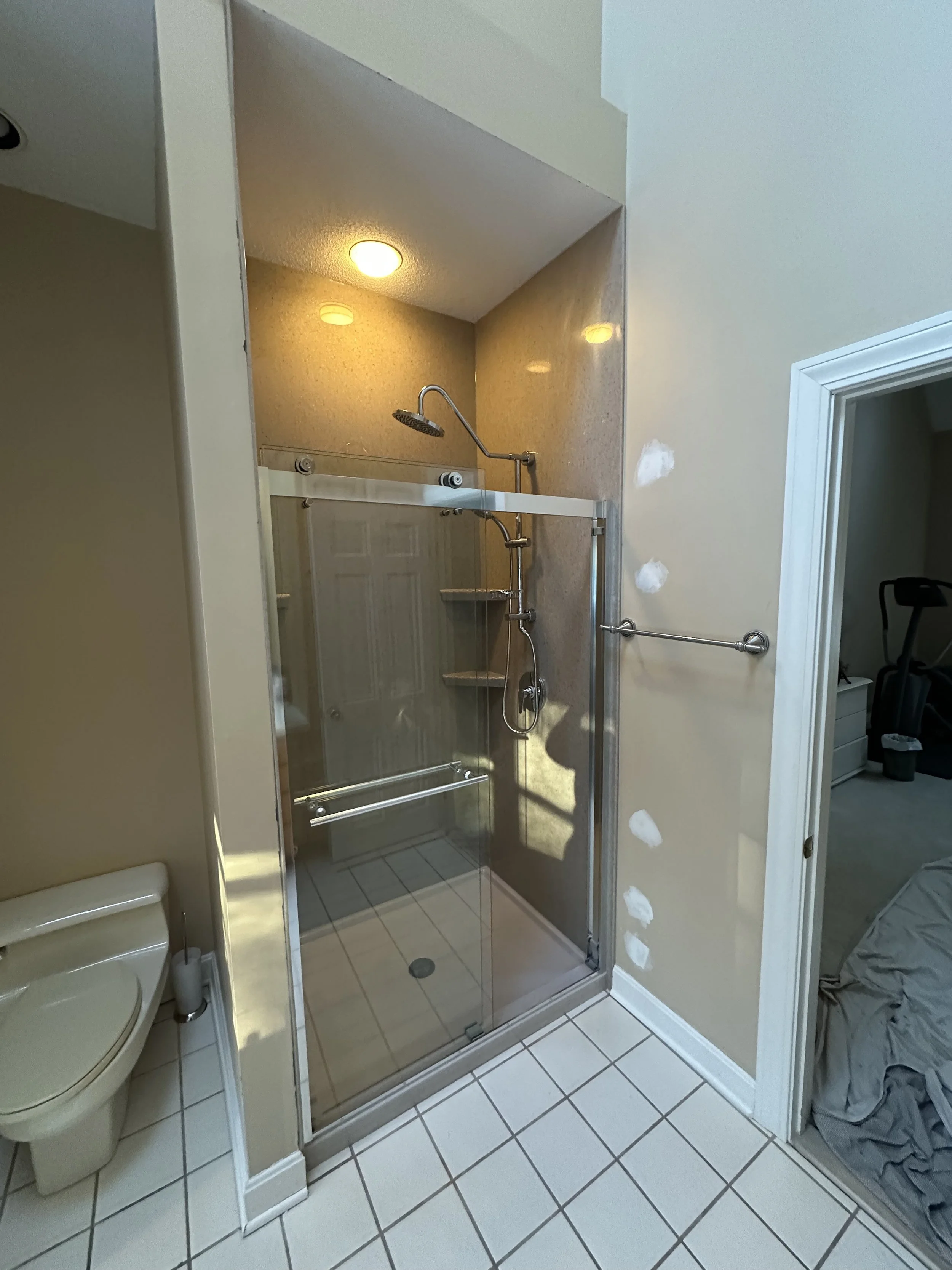 Bathroom with a glass shower enclosure, a beige wall with patches of white paint, a toilet to the left, and an open doorway leading to a bedroom with exercise equipment.