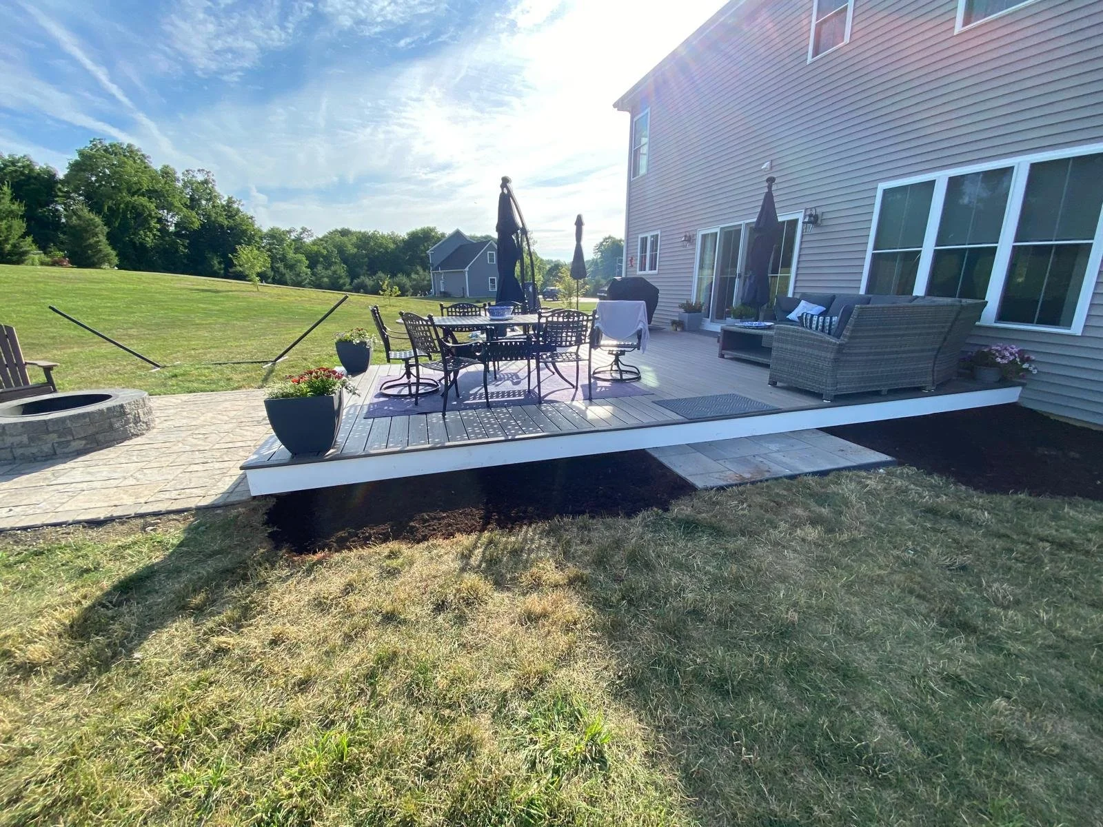 Backyard patio with outdoor furniture, including a dining table with chairs and a sofa, on a raised deck with a fire pit and planters, adjacent to a house with large windows and a sliding door, overlooking a grassy lawn and trees in the distance.