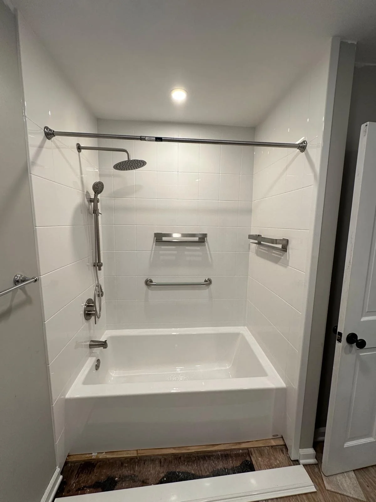 A white bathroom shower with grab bars, a shower head, and a bathtub, with white tiled walls and brown wooden flooring.
