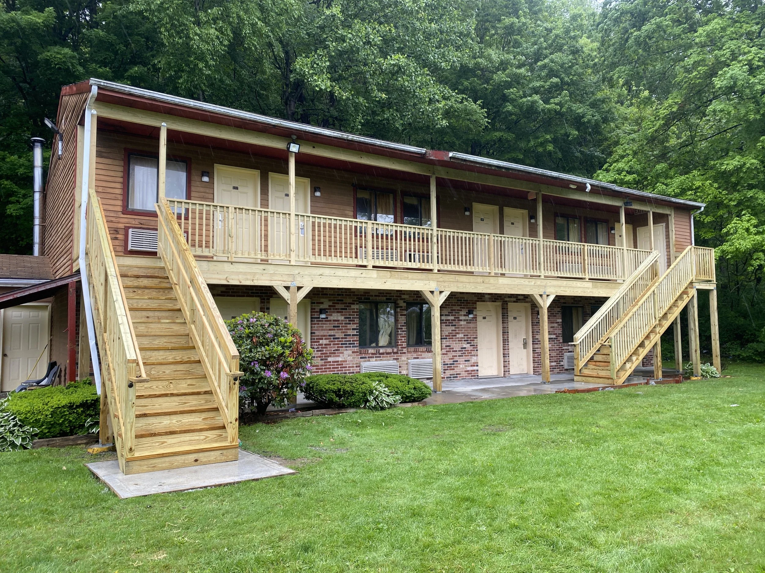 A two-story brick and wood apartment building with wooden staircases leading to the upper units, surrounded by green grass and trees.