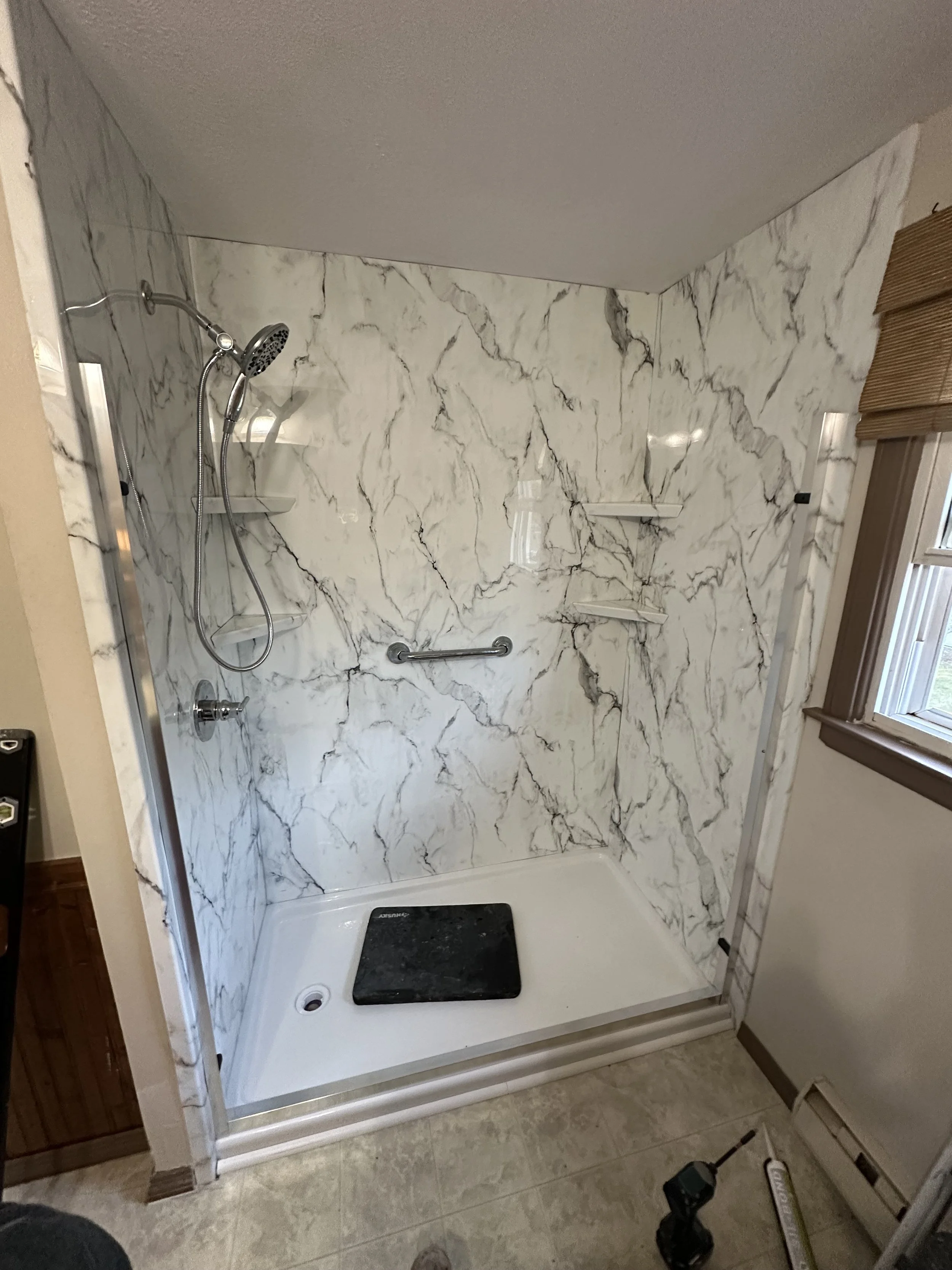 A walk-in shower with marble-patterned walls, a handheld showerhead, a grab bar, corner shelves, and a black soap dish on the white shower base. There is a window with wooden trim and partial bathroom floor visible.