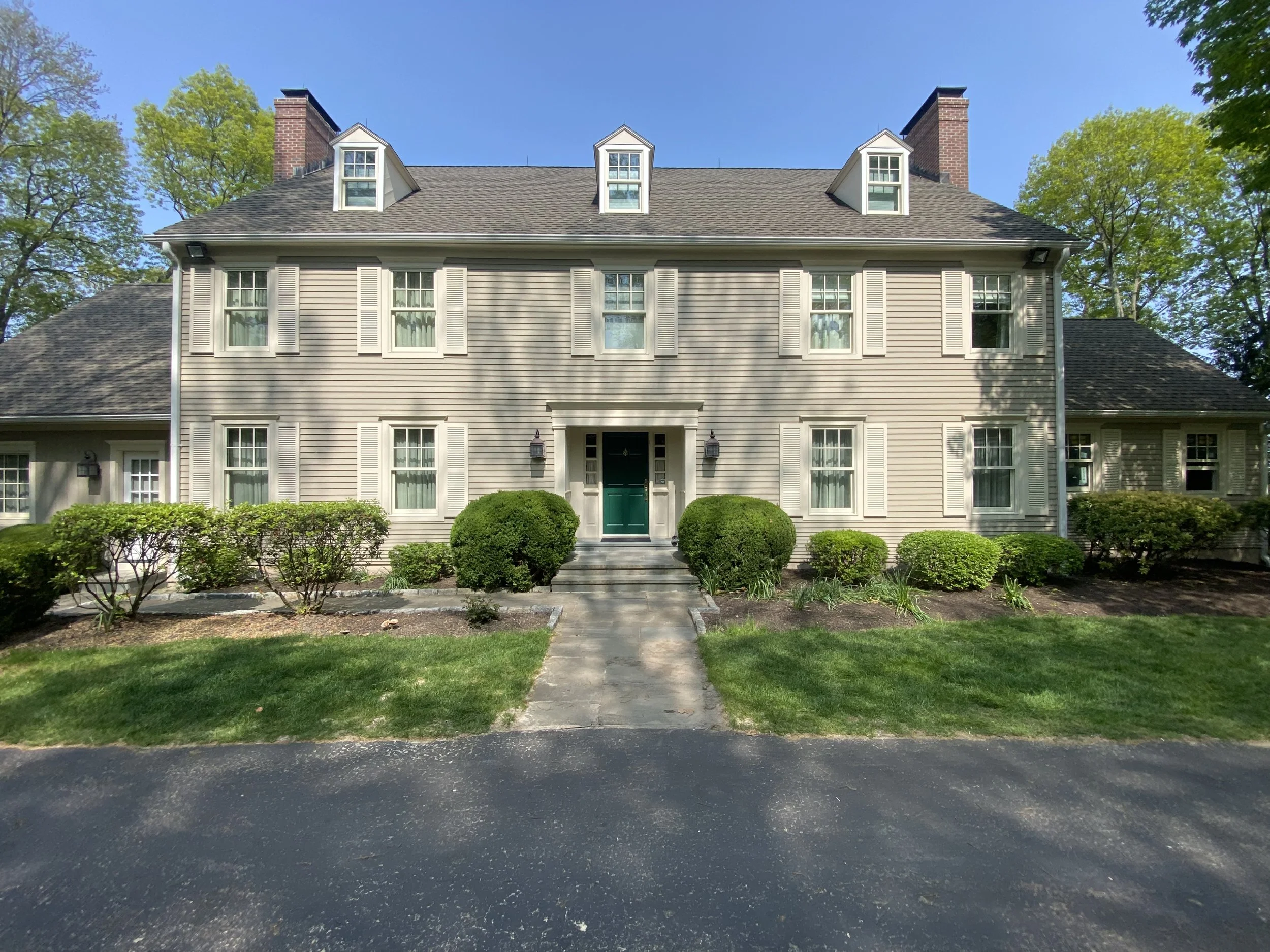 Front view of a two-story house with beige siding, white shutters, and a green front door. Well-maintained shrubbery and a paved walkway lead to the entrance, with a grassy lawn and tall trees in the background.