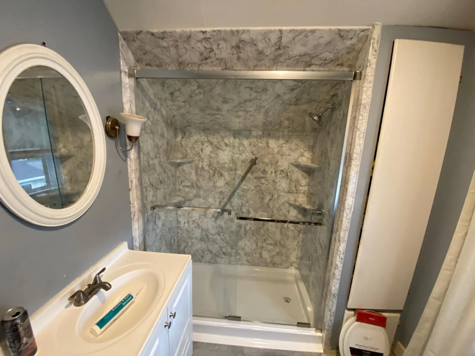 Bathroom with a white vanity sink, a round mirror, wall-mounted light fixture, a marble shower with sliding glass door, two grab bars, and a corner shelf. There is also a white cabinet and a trash can with a red lid.