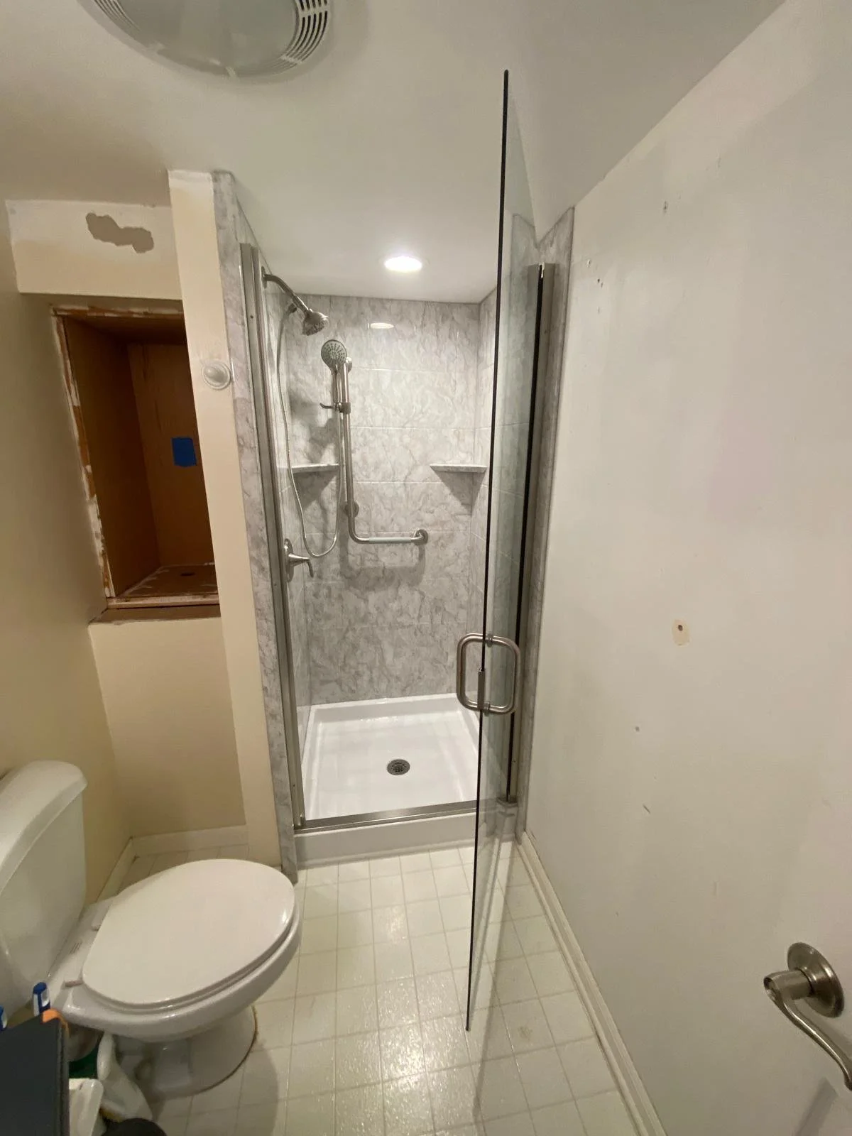 Small bathroom with a toilet on the left and a glass-enclosed shower with grab bars and built-in shelves at the back.