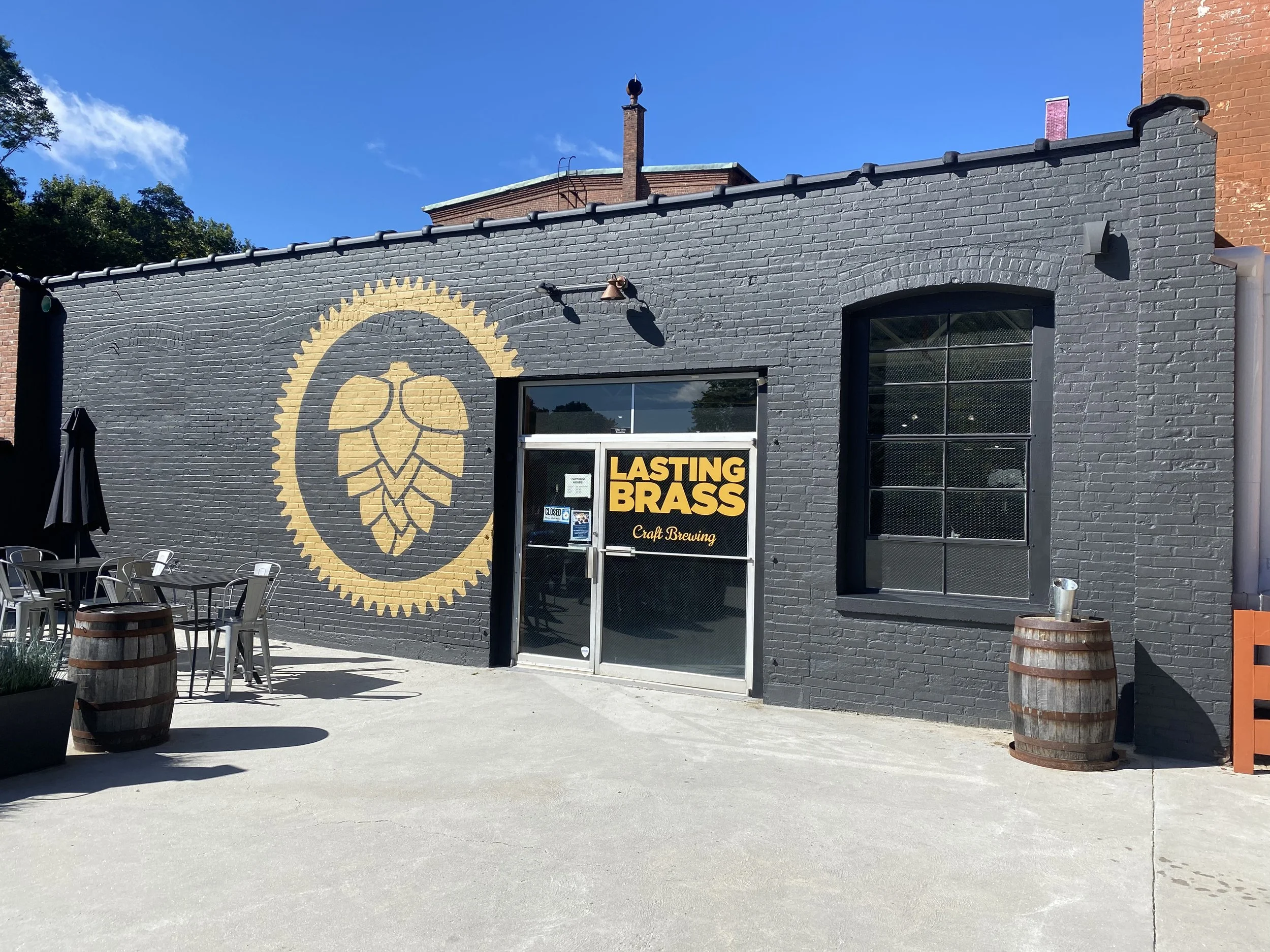 Exterior of a brick building painted dark gray with a large hop cone logo painted on the wall and a sign that reads "Lasting Brass Craft Brewing". There is outdoor seating with metal tables and chairs, wooden barrels, and umbrellas.