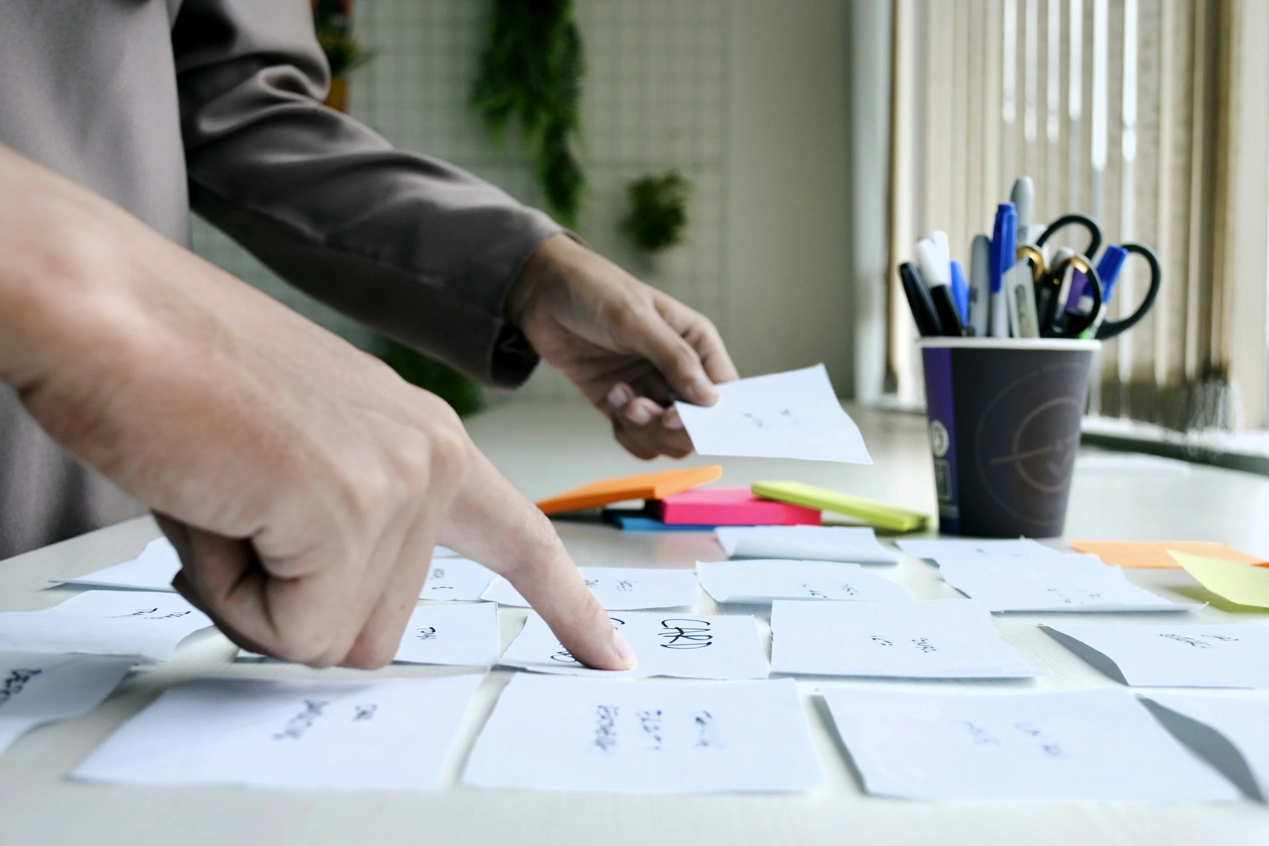 Close-up of a person's hand pointing at handwritten notes on paper, with colorful sticky notes scattered nearby and a container of various pens and markers in the background, on a desk.