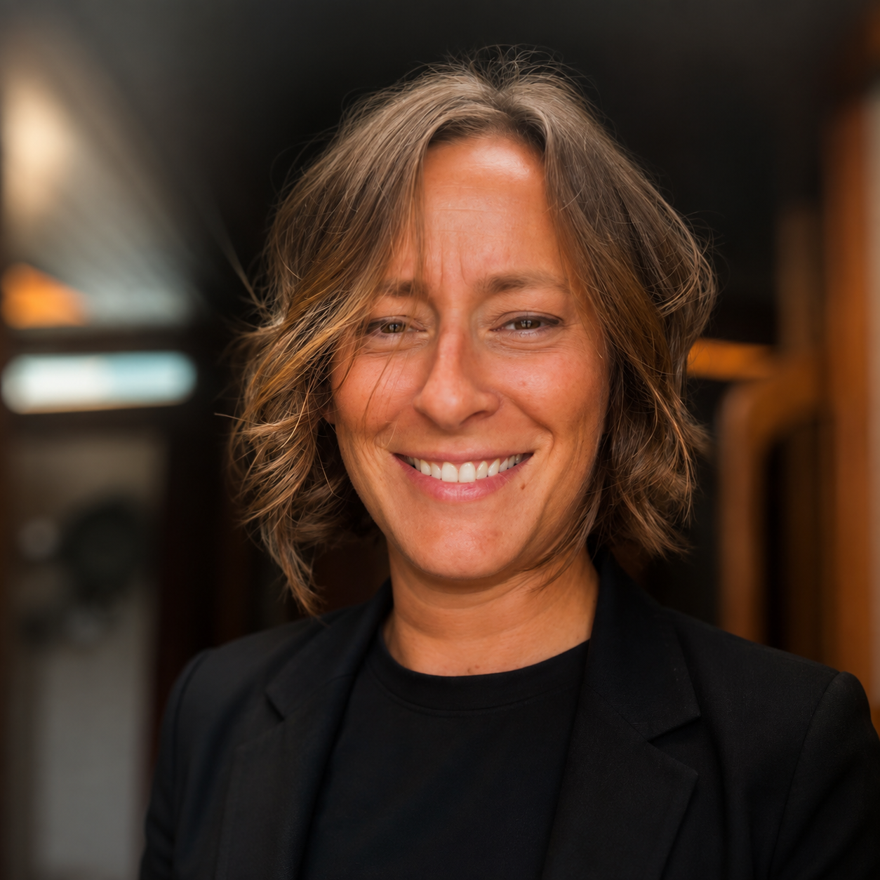 A woman with short, wavy, light brown hair smiling at the camera, wearing a black blazer and t-shirt, in an indoor setting with warm lighting.