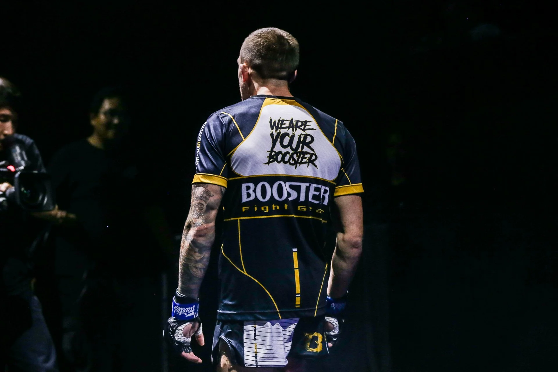 Ein männlicher Kampfsportler steht mit dem Rücken zur Kamera, trägt ein schwarzes und gelbes Fight-Shirt mit der Aufschrift 'WEARE YOUR BOOSTER' und 'BOOSTER Fight Gear', tighte Shorts, Kampfhandschuhe, und hat Tattoos auf den Armen, während er sich auf einen Kampftag vorbereitet.