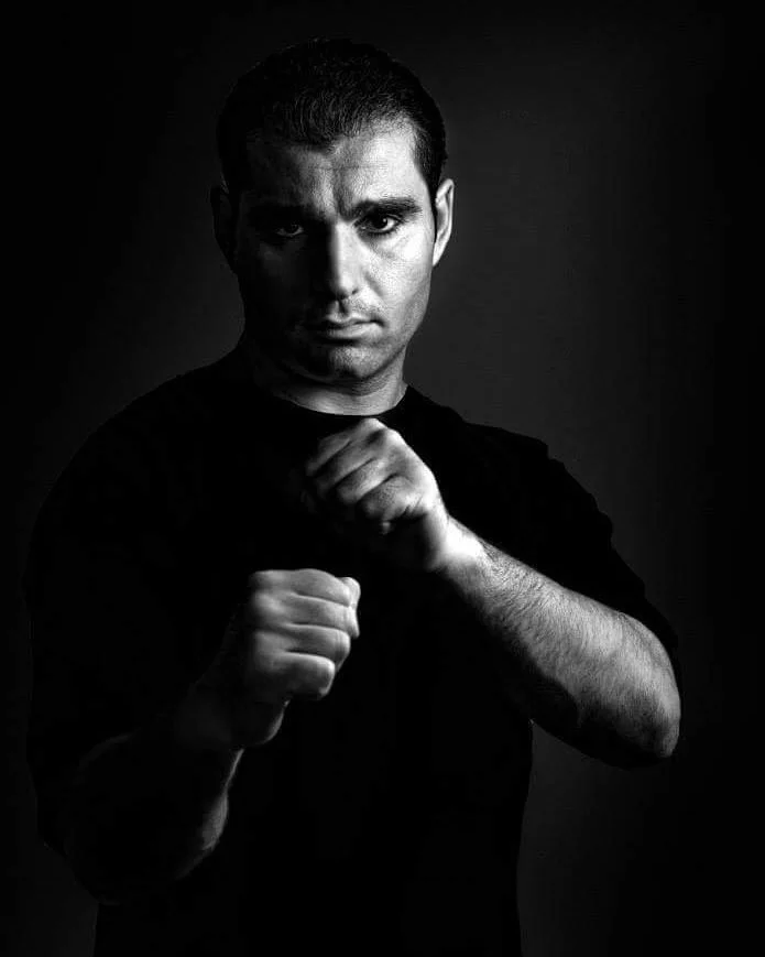 Schwarze und weiße Aufnahme eines Mannes in Kampfsporthaltung, Blick direkt in die Kamera, dunkler Hintergrund, intensiver Gesichtsausdruck.