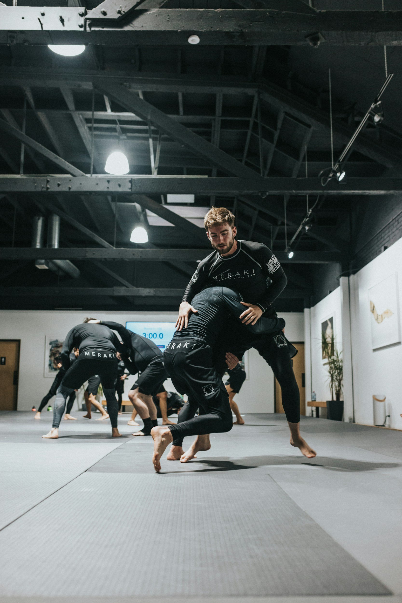 Menschen beim Brazilian Jiu-Jitsu Training in einem Gym, mehrere Personen in schwarzen Gis üben Techniken, man im Vordergrund hebt eine Person im Boden, im Hintergrund ein digitales Timer-Display