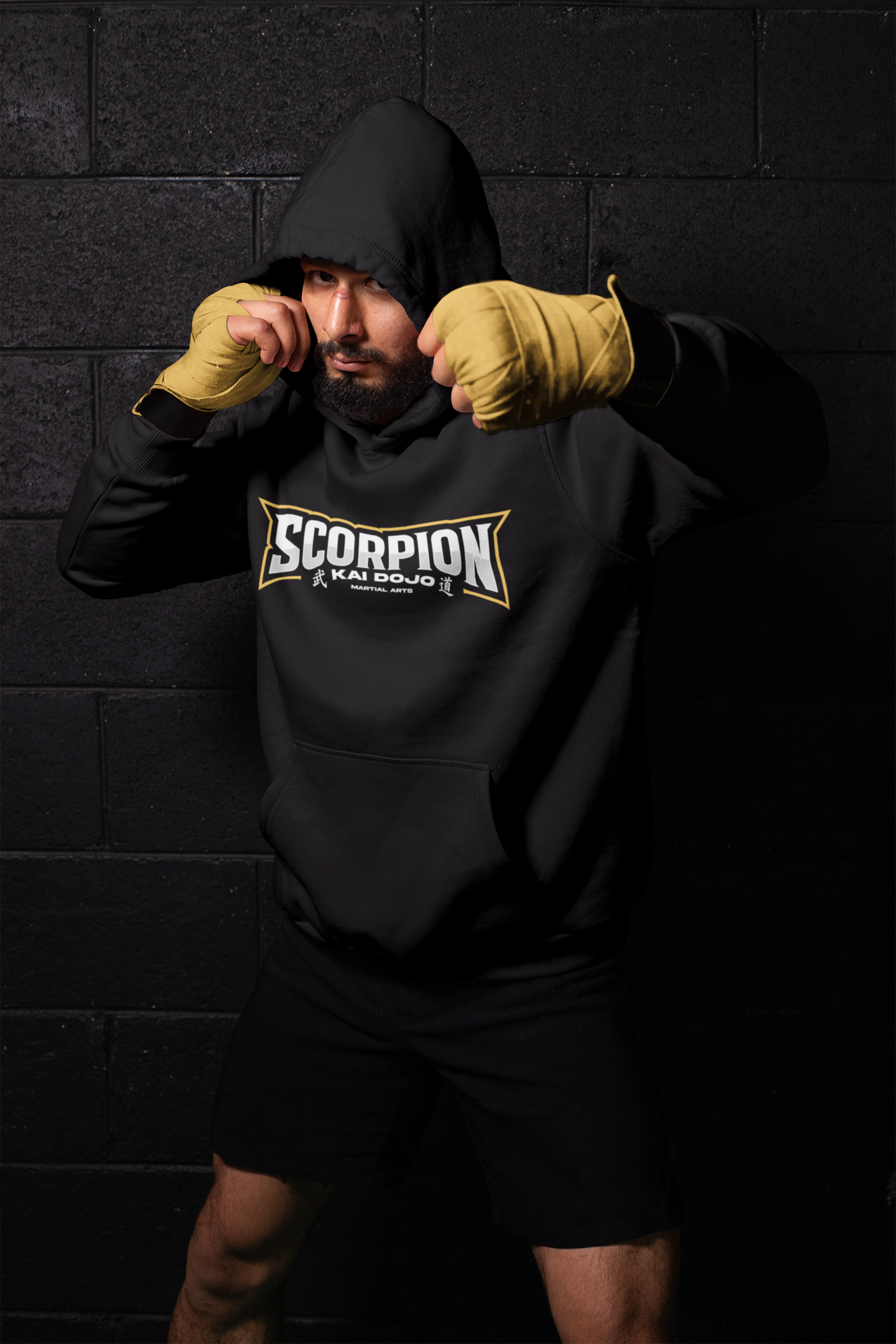 Mann in Kampfsportkleidung mit schwarzen Hoodie und gelben Boxhandschuhen in Kampfform, vor dunklem Ziegelsteinwand.