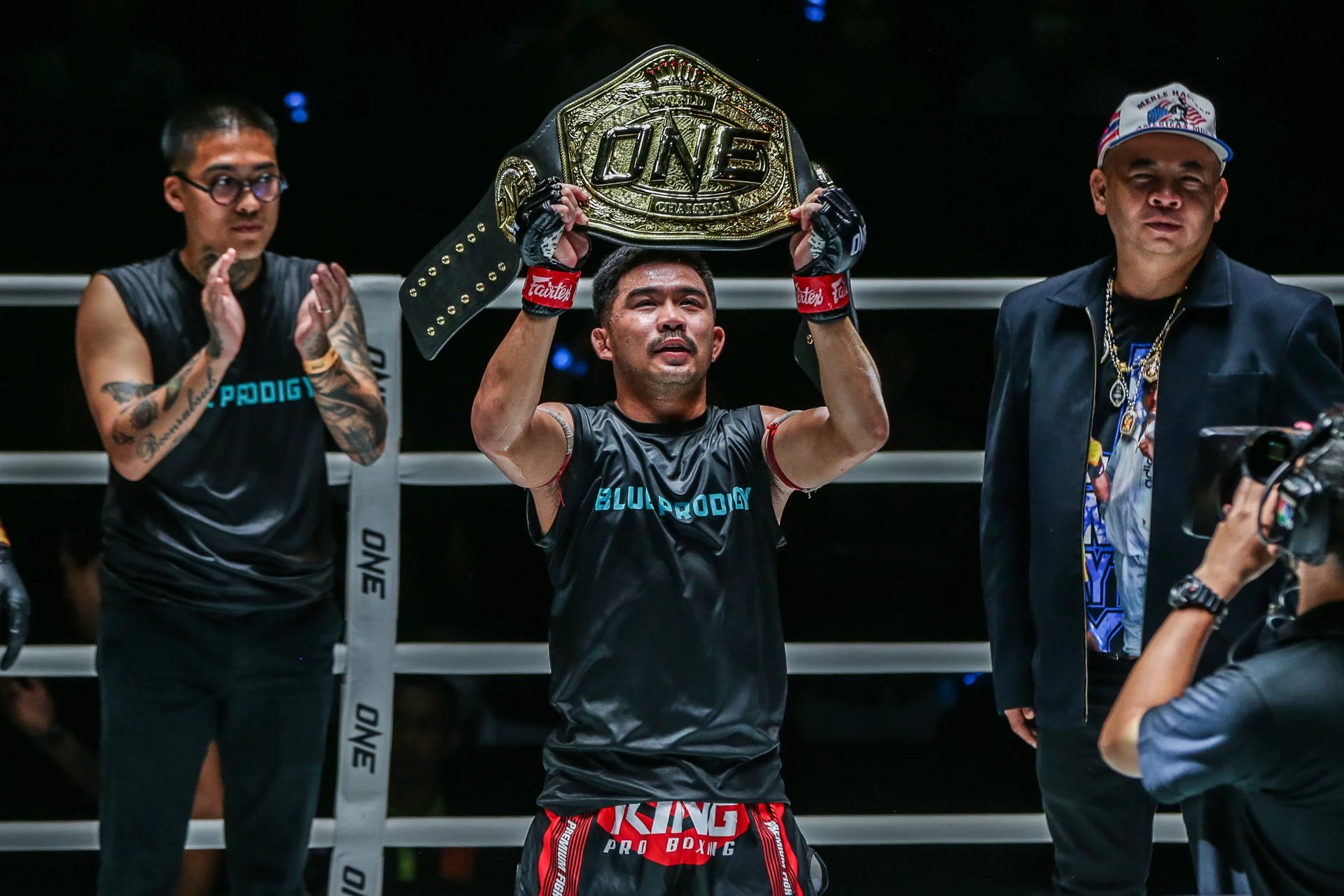 Ein Boxer hält den Meisterschaftsgürtel hoch in der Mitte des Rings, während er von zwei Personen umgeben ist, die applaudieren und ihn feiern.