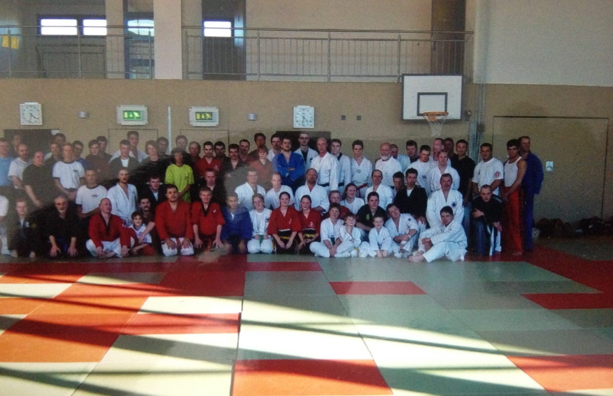Gruppenbild von Kampfsportlern auf einer Trainingsmatte in einer Turnhalle, einige in Karate- oder Judo-Gi, andere in Sportkleidung, mit einem Basketballkorb im Hintergrund.