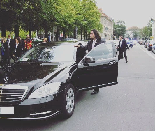 Frau steigt in einen schwarzen Mercedes auf einer Stadtstraße mit vielen Fußgängern und Bäumen.