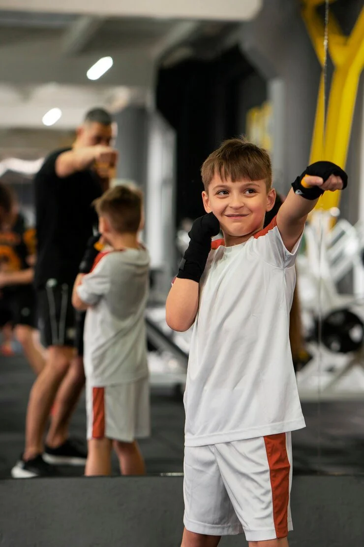 Junge im Boxanimkampf im Fitnessstudio, trägt Handschuhe und Sportkleidung, lachend, im Hintergrund andere Kinder
