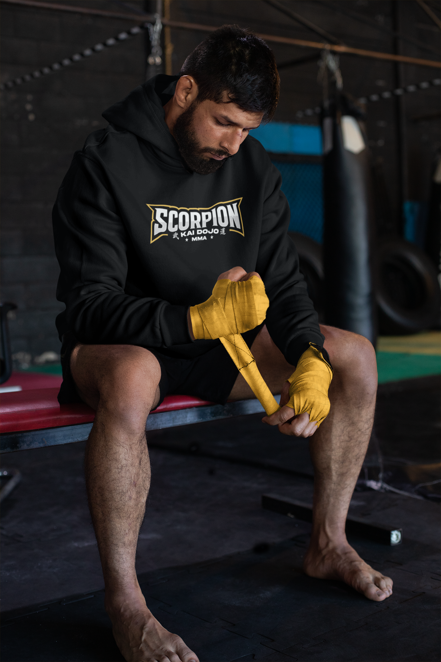 Mann sitzt auf einer Bank im Fitnessstudio, wickelt seine Hände in gelbe Handbandagen, trägt ein schwarzes Hoodie mit dem Schriftzug 'Scorpion' und hat kurze dunkle Haare und Bart.