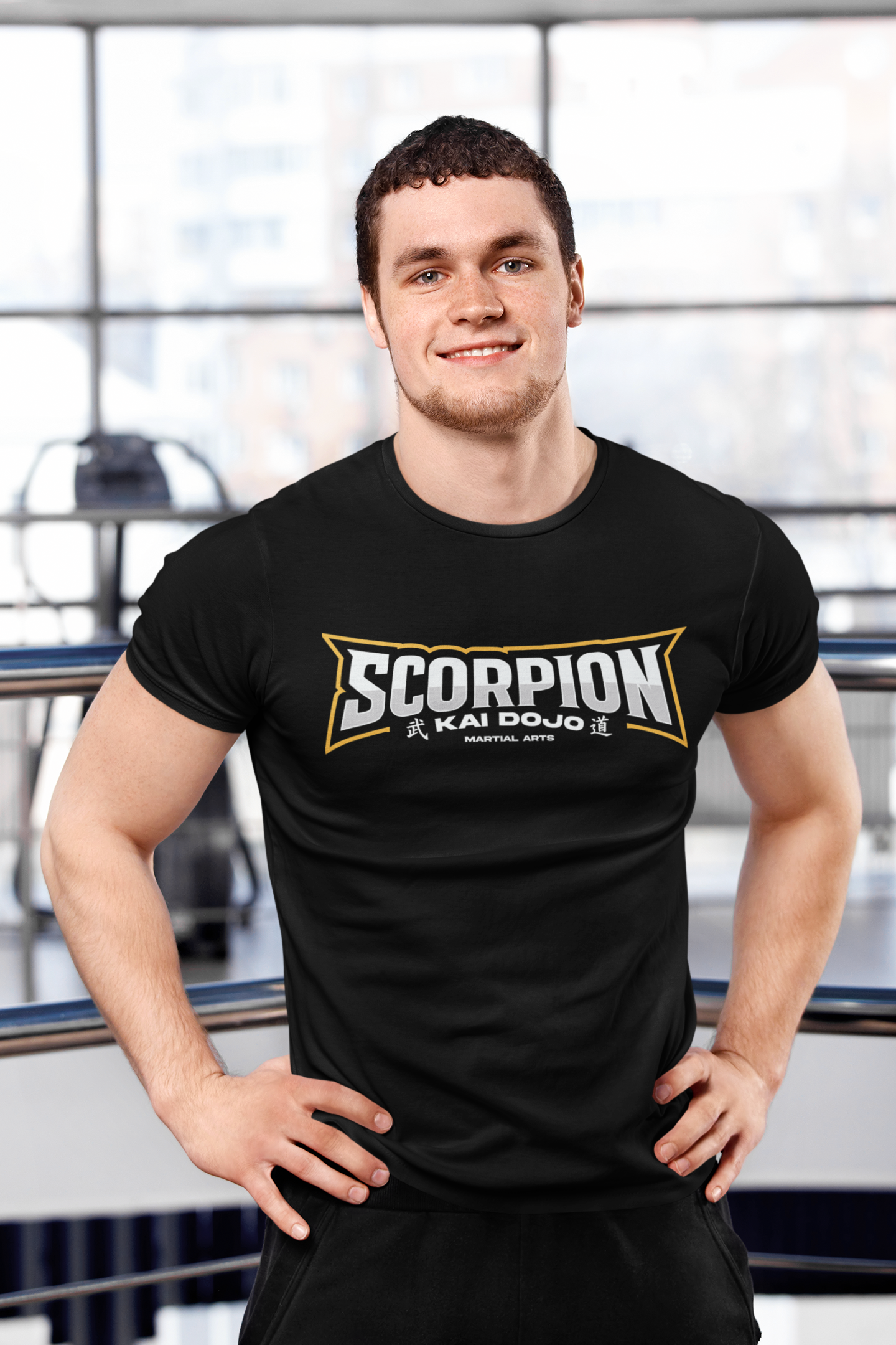 Junger Mann im Fitnessstudio trägt ein schwarzes T-Shirt mit dem Logo 'Scorpion Kai Do' und steht hinter Metallreling.