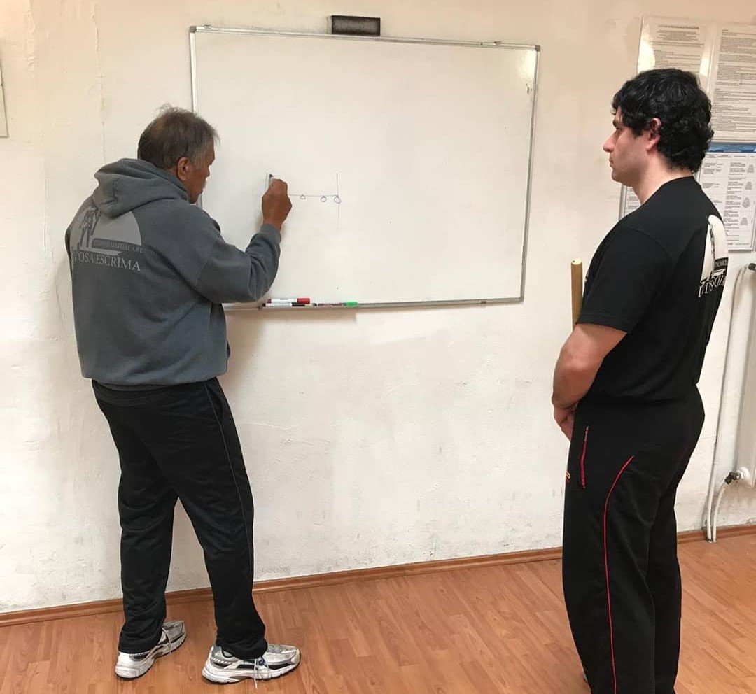 Zwei Männer stehen vor einem Whiteboard, auf dem eine Skizze mit drei kleinen Kreisen und einer Linie gezeichnet ist. Der Mann auf der linken Seite zeichnet, der Mann auf der rechten Seite beobachtet. Sie befinden sich in einem Raum mit Holzfußboden.