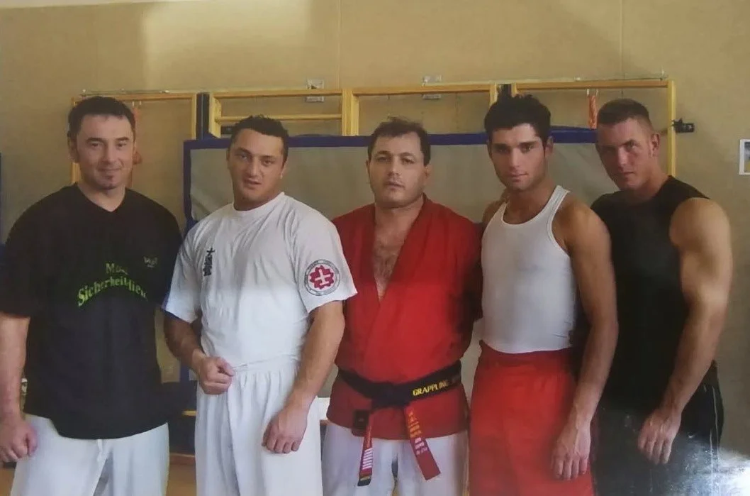 Fünf Männer in Kampfsportkleidung stehen nebeneinander in einem Trainingsraum.