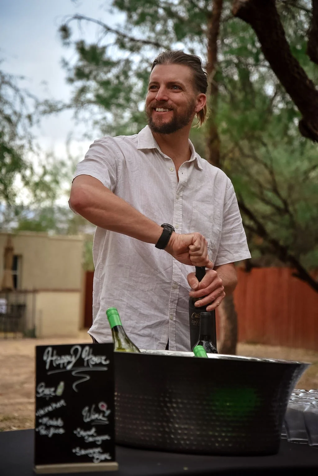 A male bartender in a white linen shirt opens a bottle of wine while smiling at an off camera guest. A happy hour sign and iced bucket of champagne and white wine sit in the foreground.