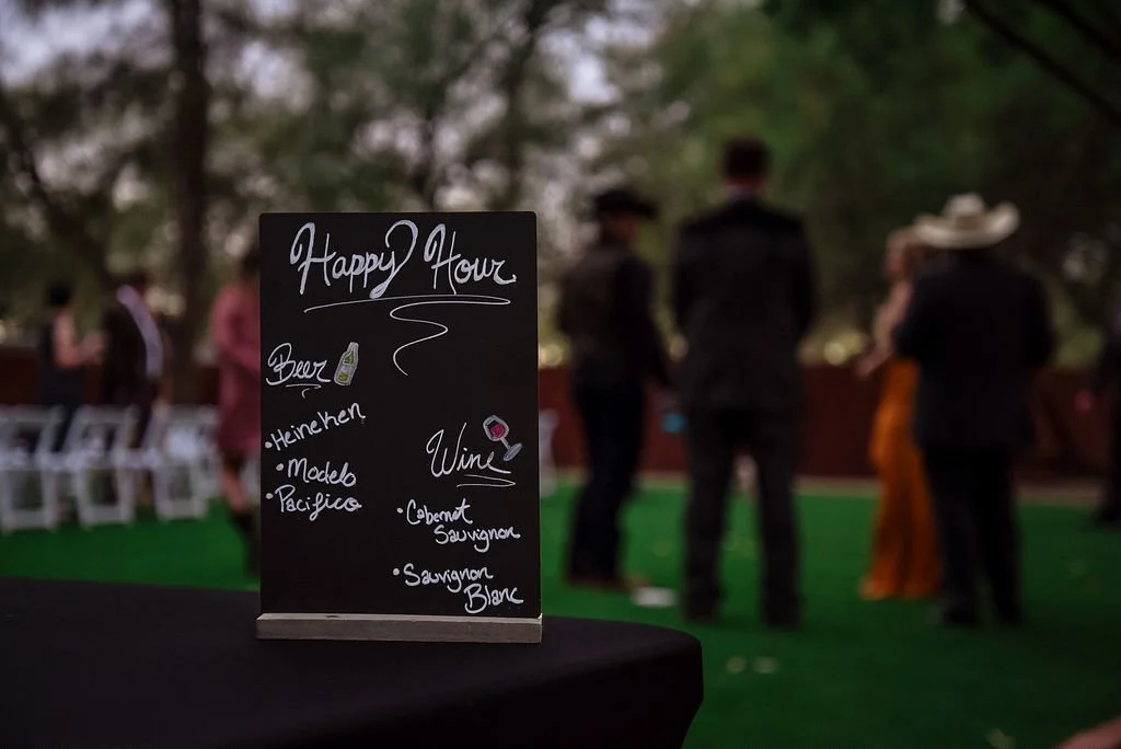 A sign featuring happy hour drinks of beer and wine sits on a table with wedding guests in the background.