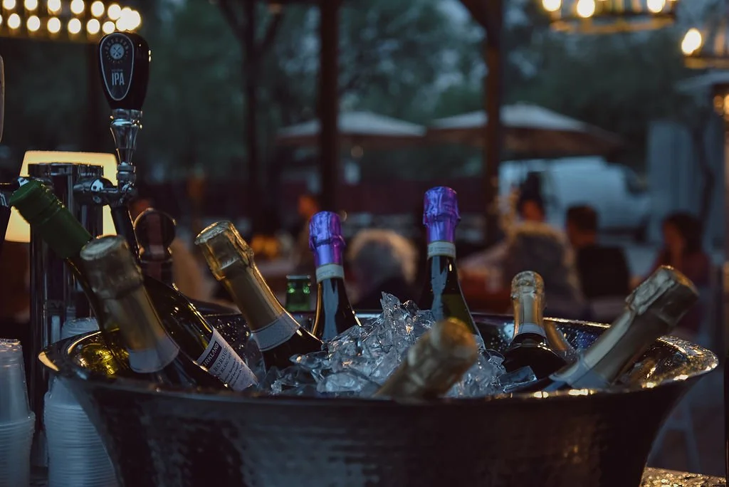 A bucket of champagne and white wine on ice sits on the counter of a bar at an outdoor wedding while guests enjoy their food in the background.
