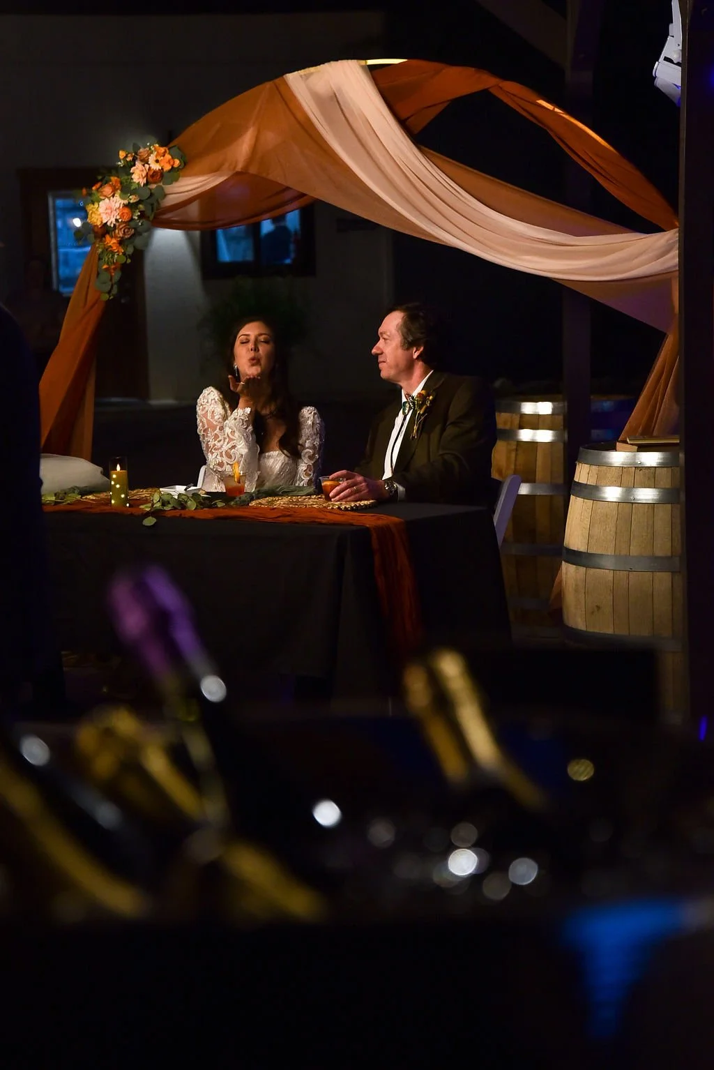 A bride blows a kiss to the camera while she sits next to her new husband at a table at their Arizona wedding. There are wine barrels in the background.