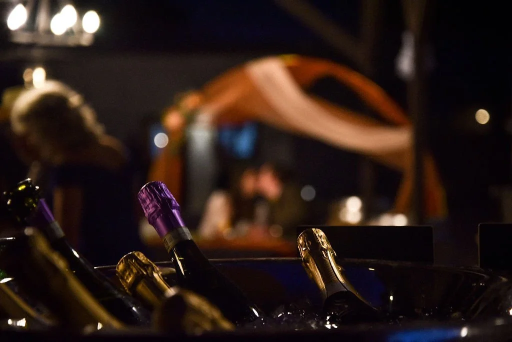 The necks of champagne bottles are in focus in the foreground of the image while a newlywed couple shares a kiss in the background.