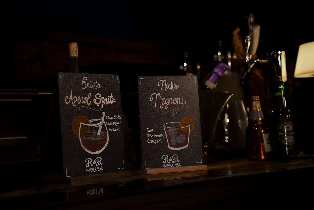 Small chalkboard signs show drawings of an aperol spritz and a negroni and their ingredients. The signs sit on the counter of an outdoor bar next to bottles of beer and wine.